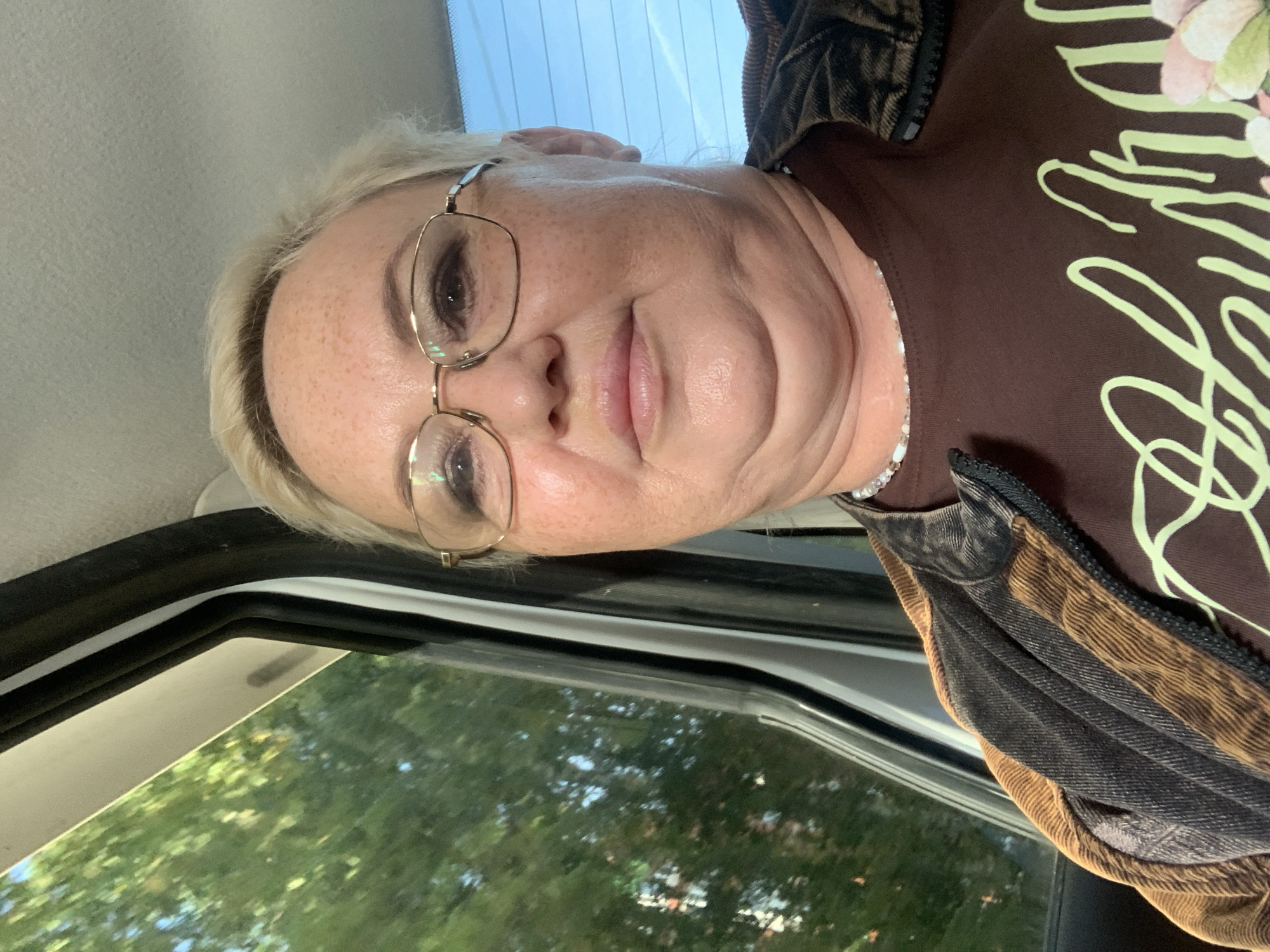 A middle-aged woman with short blonde hair and freckles is looking into the camera from the passenger seat of a vehicle. She is wearing glasses, a brown t-shirt with a light green graphic, and a brown and black jacket. The background shows trees and foliage through the car window, suggesting an outdoor environment, possibly in Chaikovsky, Russia. The lighting is natural, indicating daytime.