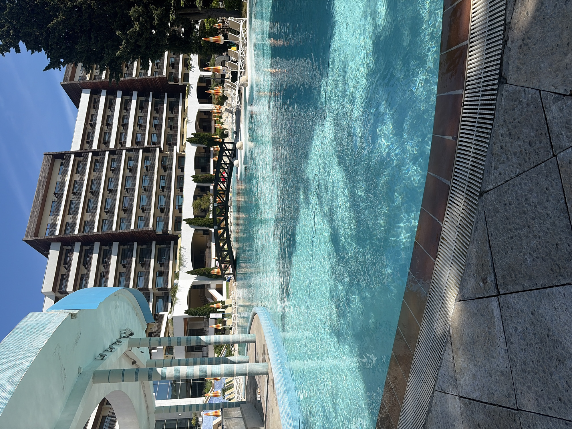 The image, oriented vertically, captures a vibrant outdoor swimming pool scene at a resort in Obrochishte, Bulgaria, under a clear blue sky. A large, inviting swimming pool with clear, rippling blue water occupies the right side of the frame, bordered by a dark tiled gutter system featuring a metallic grate, adjacent to gray, textured paving stones. The pool's edge is also detailed with blue mosaic tiles in the foreground.
On the left, a multi-story hotel or resort building stands prominently, showcasing a modern architectural style with numerous balconies and alternating white and brown-wood panel facades. Along the poolside, several white lounge chairs are arranged, accompanied by distinctive orange and white striped sun umbrellas, suggesting an area for relaxation. Small, green manicured trees and shrubs are visible near the building's base.
In the foreground, a striking light blue and white tiled archway structure with several cylindrical pillars and mounted spotlights adds a decorative element to the scene. Further back, a dark, arched bridge-like structure with spherical white lights spans a section of the pool. A large, dark green tree frames the upper left corner, contrasting with the bright sky. Indistinct figures are visible in the pool, indicating people are swimming or enjoying the water. The strong sunlight and clear shadows confirm it is daytime. No visible text is present in the image.