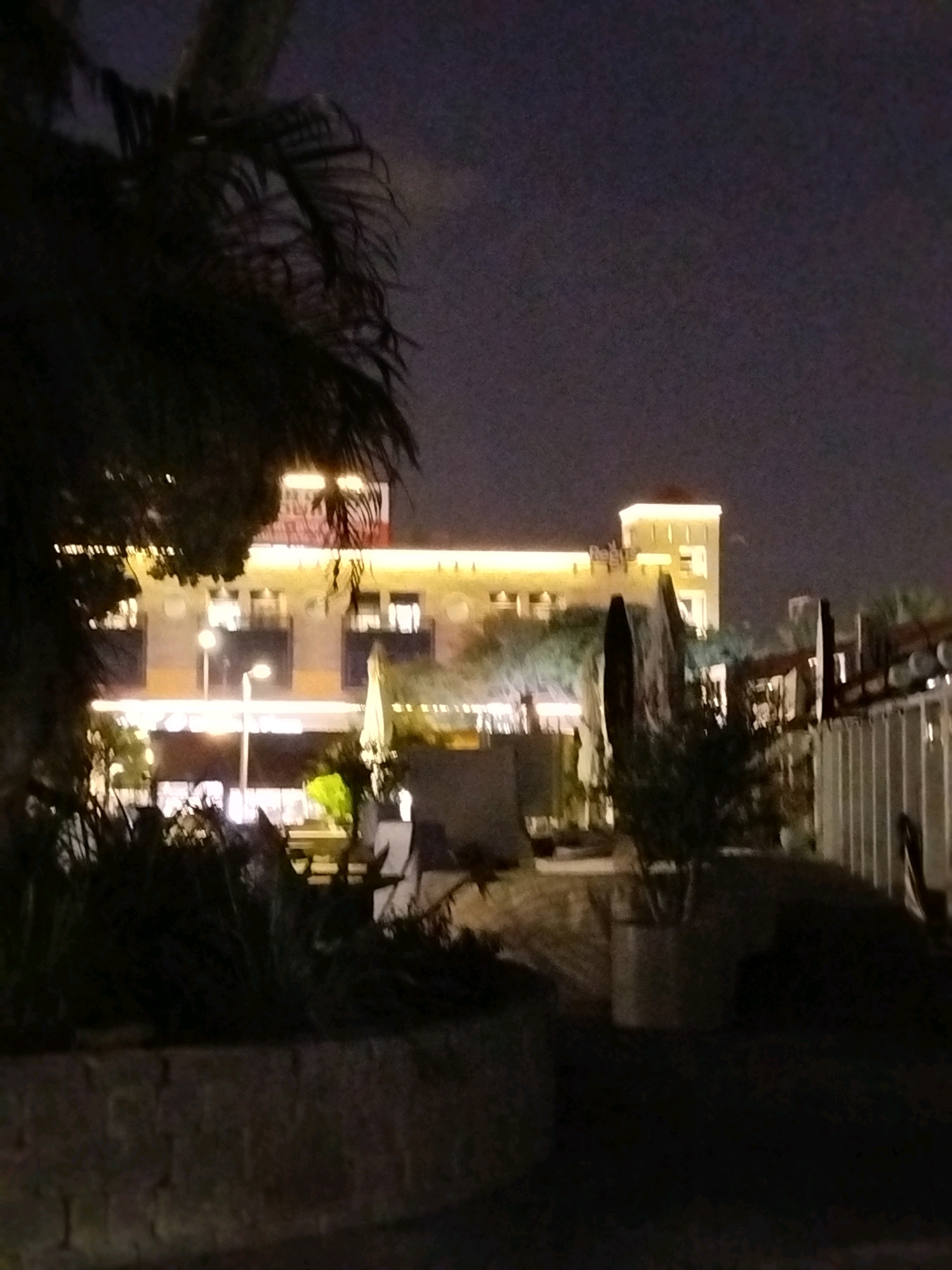 This image depicts a nighttime scene in Ashkelon, Israel, likely at an outdoor dining or entertainment area. In the background, a multi-story building is illuminated with warm lighting. The building features several floors with visible windows and balconies. A prominent tower-like structure with a light-colored roof protrudes from the building. In front of the building, there are outdoor seating areas with tables and chairs, interspersed with greenery and trees. The foreground is dark, with shadows and a low stone wall or planter. A palm tree is visible in the upper left portion of the frame, its fronds silhouetted against the dark sky. The overall atmosphere suggests a lively evening ambiance in a commercial district of Ashkelon.