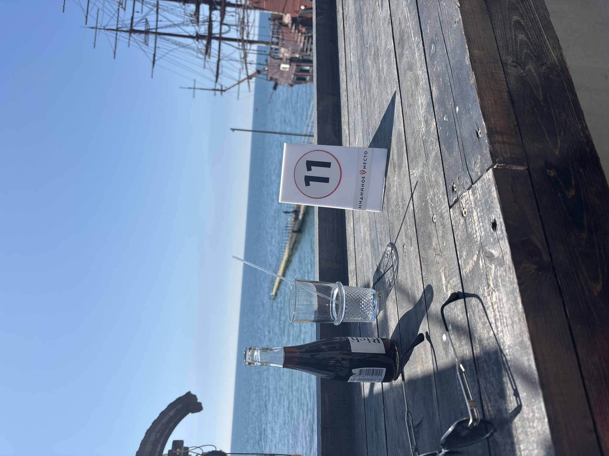 On a sunny day in Sochi, Russia, a table on a wooden deck overlooks the sea. A table number stand displays "11". A bottle of dark liquid lies next to a glass with a straw, and sunglasses rest on the table. A large ship can be seen in the background.