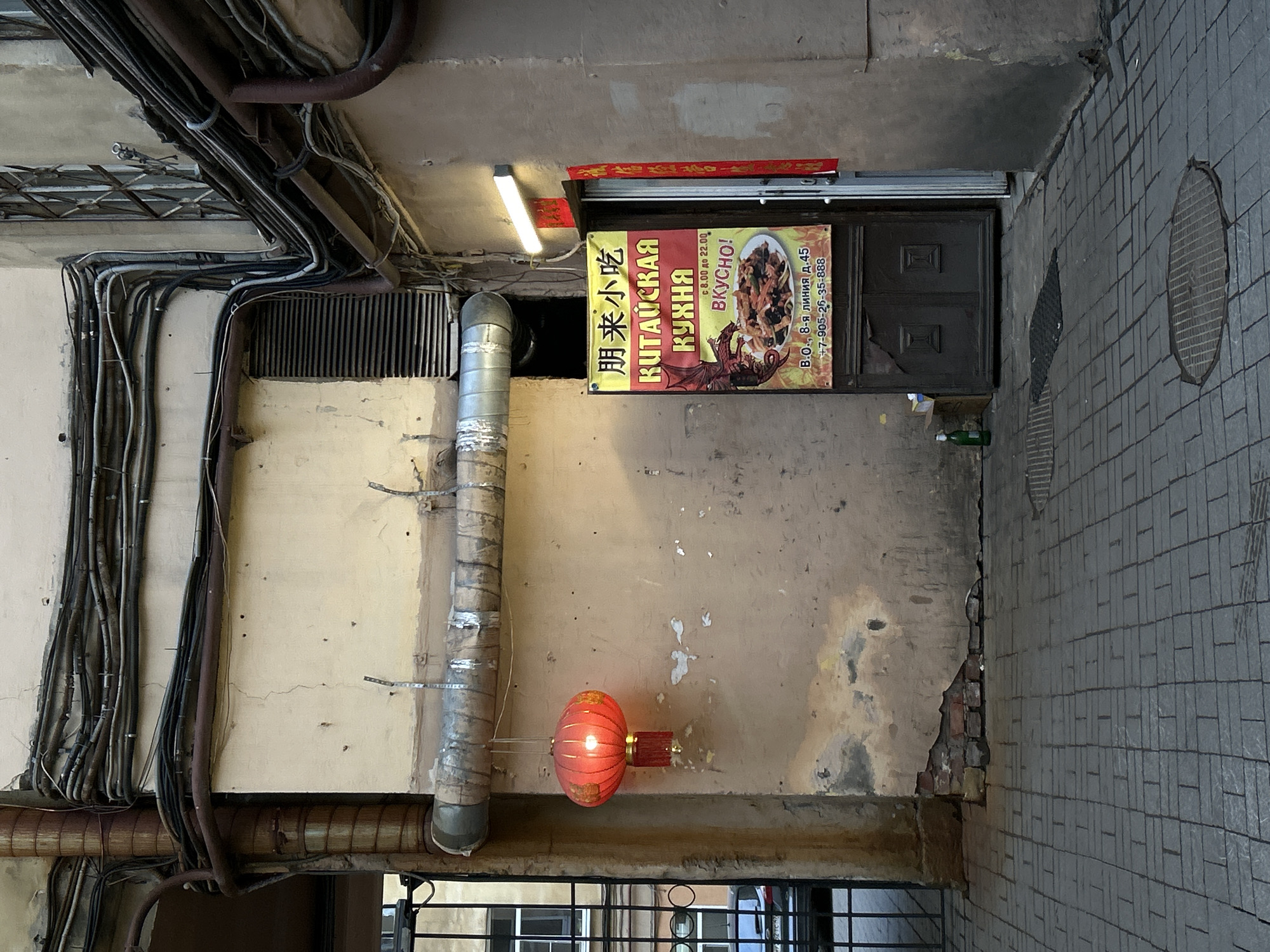 The image, oriented vertically, depicts a narrow, somewhat unkempt urban passage or courtyard. The scene is dominated by a wall on the left, extensively covered with an intricate network of exposed pipes and electrical conduits running along its surface and ceiling. The wall itself shows signs of age, with peeling paint and exposed concrete, appearing largely light beige or cream-colored.
At the center, affixed to a wall above a dark wooden double door, is a brightly colored sign for a Chinese restaurant. Below this, a red, illuminated Chinese lantern hangs from a bracket, adding a distinct cultural element. To the right, the ground is paved with grey rectangular tiles, and a circular manhole cover is visible. The overall impression is one of an older, utilitarian urban space.
No people are visible, and no specific activity is taking place, although the sign indicates a commercial establishment. The lighting suggests daytime, possibly overcast, or within a shadowed courtyard.
Visible text on the sign includes:
* Chinese characters: "朋来小吃" (Péng lái xiǎochī), translating to "Peng Lai Snacks" or "Friendship Snacks."
* Russian text: "КИТАЙСКАЯ КУХНЯ" (Kitaïskaya Kukhnya), meaning "Chinese Cuisine."
* "ВКУСНО!" (Vkusno!), meaning "Delicious!"
* Operating hours: "с 8.00 до 22.00" (from 8:00 to 22:00).
* Address: "В.О., 8-я линия д.45" (V.O., 8th Line, house 45), an address in Saint Petersburg.
* Phone number: "+7-905-26-35-888".
The sign also features a graphic of a dragon and a plate of stir-fried food. A small red decorative banner with Chinese characters is placed above the sign.