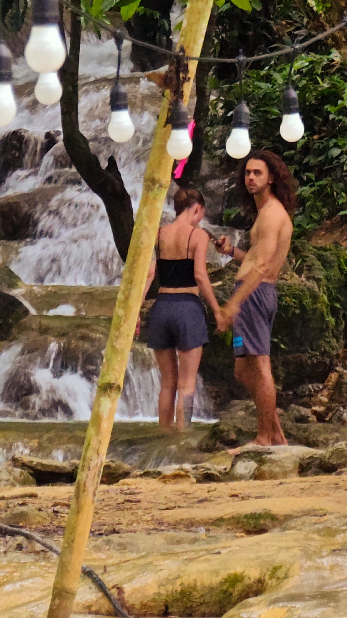 In Cổ Lũng Commune, Vietnam, a young couple stands near a waterfall. The woman, facing away from the camera, wears a black crop top and dark blue shorts. She holds a smartphone in her right hand. The man, with long, dark, curly hair, faces the camera and is shirtless. He wears dark blue shorts with a light blue accent. His left hand is held by the woman. Both are barefoot. They are positioned on rocky terrain adjacent to the cascading water. Overhead, a string of bare lightbulbs hangs from branches and a bamboo pole. Lush green foliage and a brown tree trunk are visible behind them. The foreground is dominated by wet, yellowish rocks and shallow water.