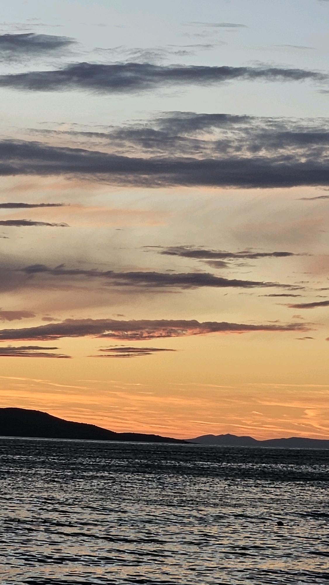The image captures a serene twilight scene over the Adriatic Sea, likely near Općina Tučepi, Croatia. Silhouetted against the vibrant orange and pink hues of the sunset are the dark outlines of coastal hills and distant mountains. The sea is a deep, textured grey, reflecting the colors of the sky with a rippling surface. Wispy clouds, ranging in color from pale blue to dark grey, stretch across the horizon, adding depth and drama to the sky. The overall impression is one of tranquil natural beauty at the end of the day.
