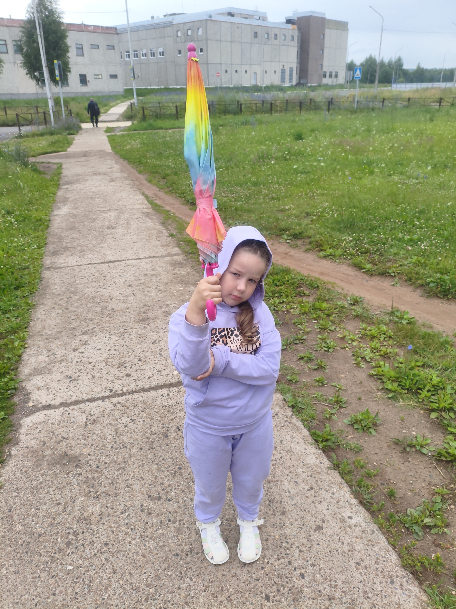 A young girl stands on a paved path, holding a closed, rainbow-colored umbrella. She is wearing a light purple hooded sweatshirt and matching pants, with white sandals. Her arms are crossed, and she is looking towards the camera with a slight frown. Behind her, a person in dark clothing walks away on the same path, leading towards a long, light-colored building with multiple windows. The building appears to be institutional or industrial. The path is bordered by grass and some dirt patches with sparse vegetation. The sky is overcast. This scene is likely located in the city of Markovsky, Russia, given the context of the building and the surrounding environment.