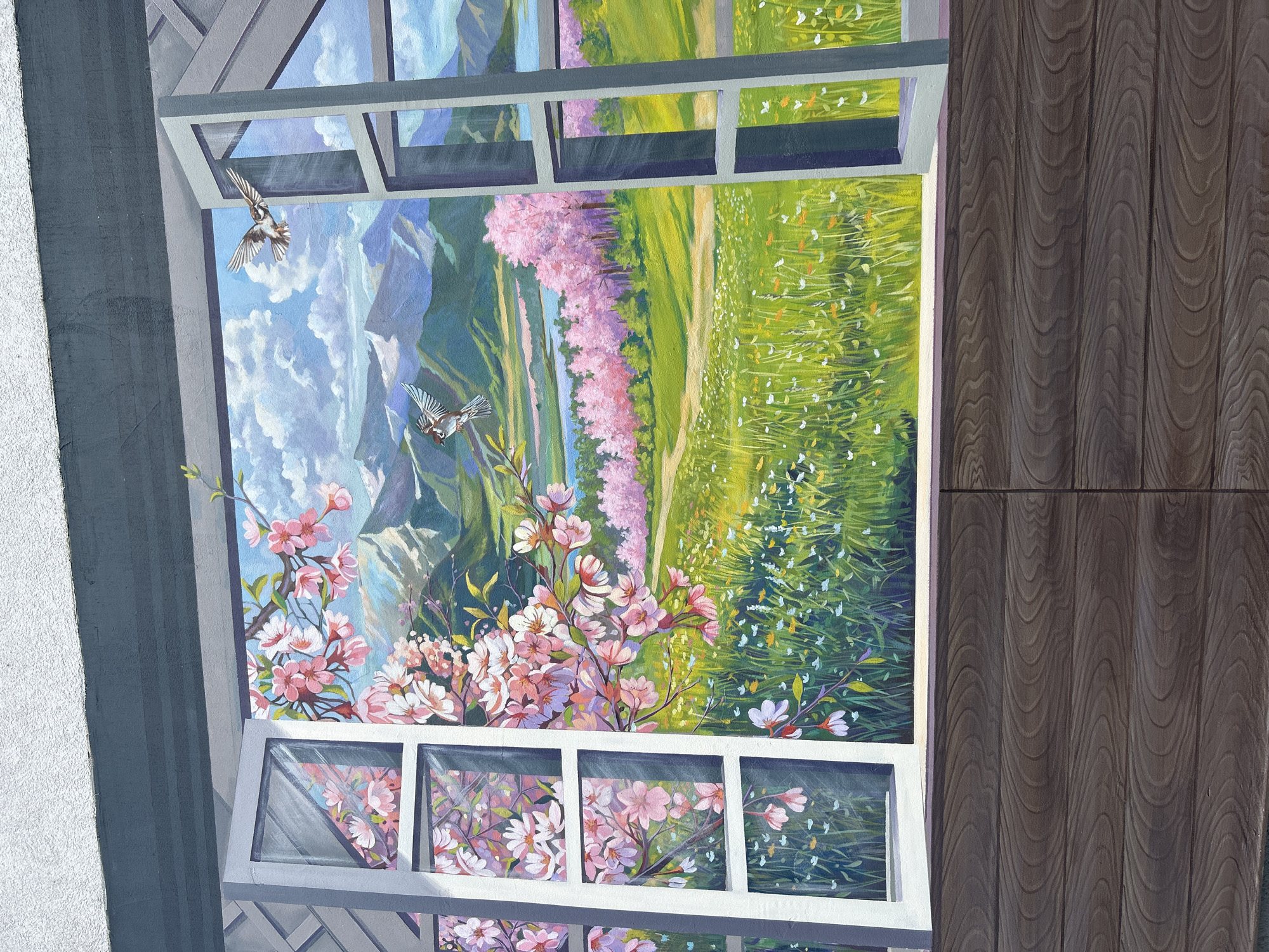 A mural depicts a view of mountains, meadow, and cherry blossoms through a white, gridded window. Two birds fly through the sky. The mural appears to be painted on an exterior wall. To the right is a wall that is painted to look like wooden planks. The location is Unknown, Unknown.