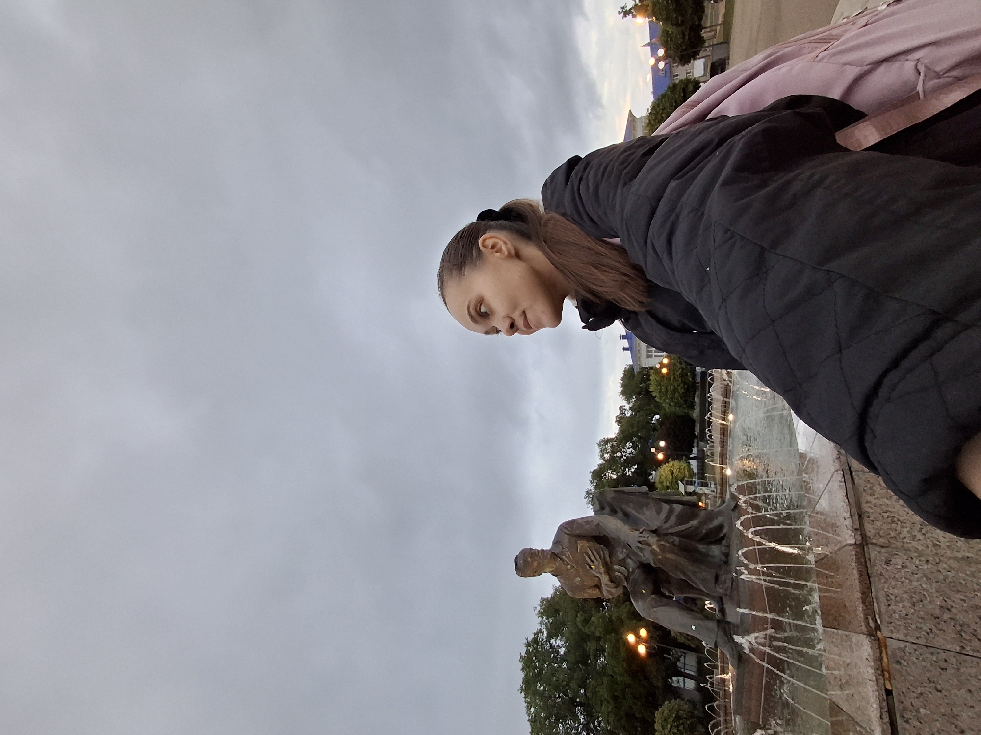 This image captures a young woman in the foreground with the Pyotr Ilyich Tchaikovsky monument and fountain in the midground, located in Chaikovsky, Russia, under an overcast sky during dusk.
In the lower right quadrant, a woman with dark hair pulled into a ponytail and wearing a black quilted jacket is depicted from a low, upward-looking angle. She has a slight smile and is gazing towards her left. A pink backpack or garment is visible on her right side. Her right arm, covered by the jacket, is extended forward, suggesting she is taking a selfie.
To the left of the woman, the bronze statue of a seated man, identified as Pyotr Ilyich Tchaikovsky, occupies the center-left midground. The statue features the man in period attire, holding an object in his left hand. It is situated within a multi-tiered fountain where numerous thin streams of water spray upwards from a light-colored stone basin.
The background consists of dense green foliage from trees, interspersed with several illuminated streetlights that emit a warm, orange glow. Beyond the trees and lights, faint architectural structures with dark blue roofs are discernible against the horizon. The sky is uniformly gray and cloudy, indicating an overcast condition or late evening. The ground level in the immediate vicinity of the fountain and woman appears to be a paved public square.