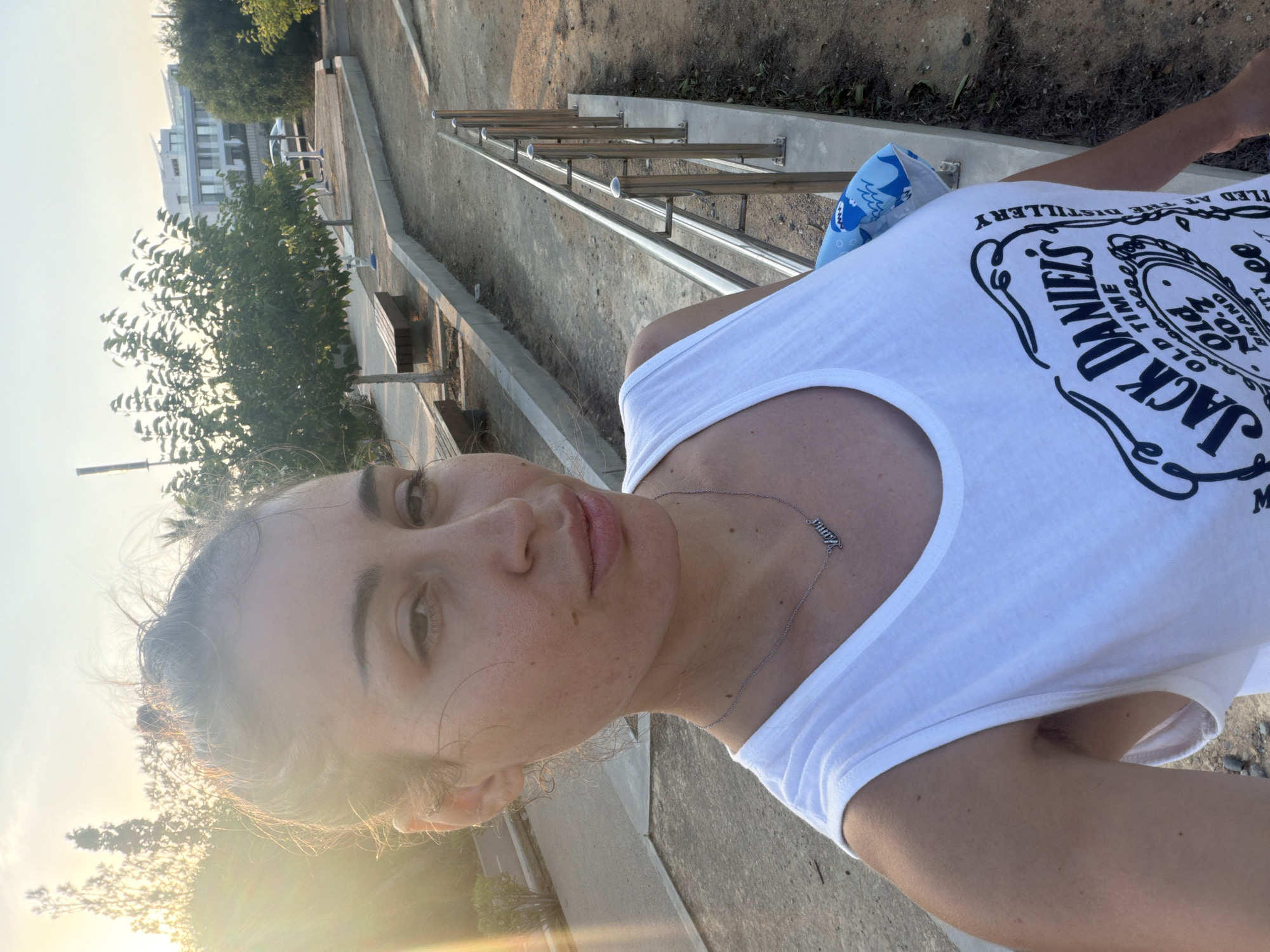 In Meneou, Cyprus, a woman with her hair in a bun takes a selfie outdoors during golden hour. She wears a white tank top with a Jack Daniel's logo. A silver necklace with the word "Mama" is visible. The background shows a paved pathway with a metal railing, trees, benches, and modern buildings. The sun casts a warm glow on the scene.
