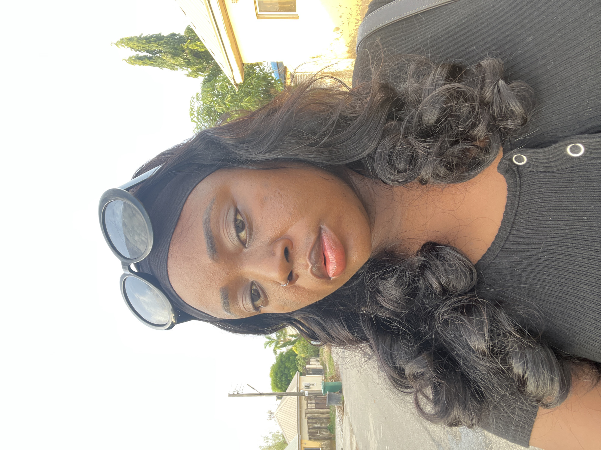 A medium shot of a woman in an outdoor setting. She is wearing a black top and has dark, curly hair, adorned with a black headband and sunglasses resting on top of her head. She has dark skin, and the photograph is taken at a close angle, highlighting her facial features, including a nose ring.
The backdrop shows a typical street scene, consisting of buildings, vegetation, and utility poles. The houses in the background appear to be single-story structures, with one painted in a combination of light and dark tones, and another with a distinct light color and a dark roof. The sky is bright and clear, suggesting a sunny day.
The location is possibly a residential area within the city of Abuja, Nigeria, judging by the architectural style of the buildings and the typical street infrastructure.