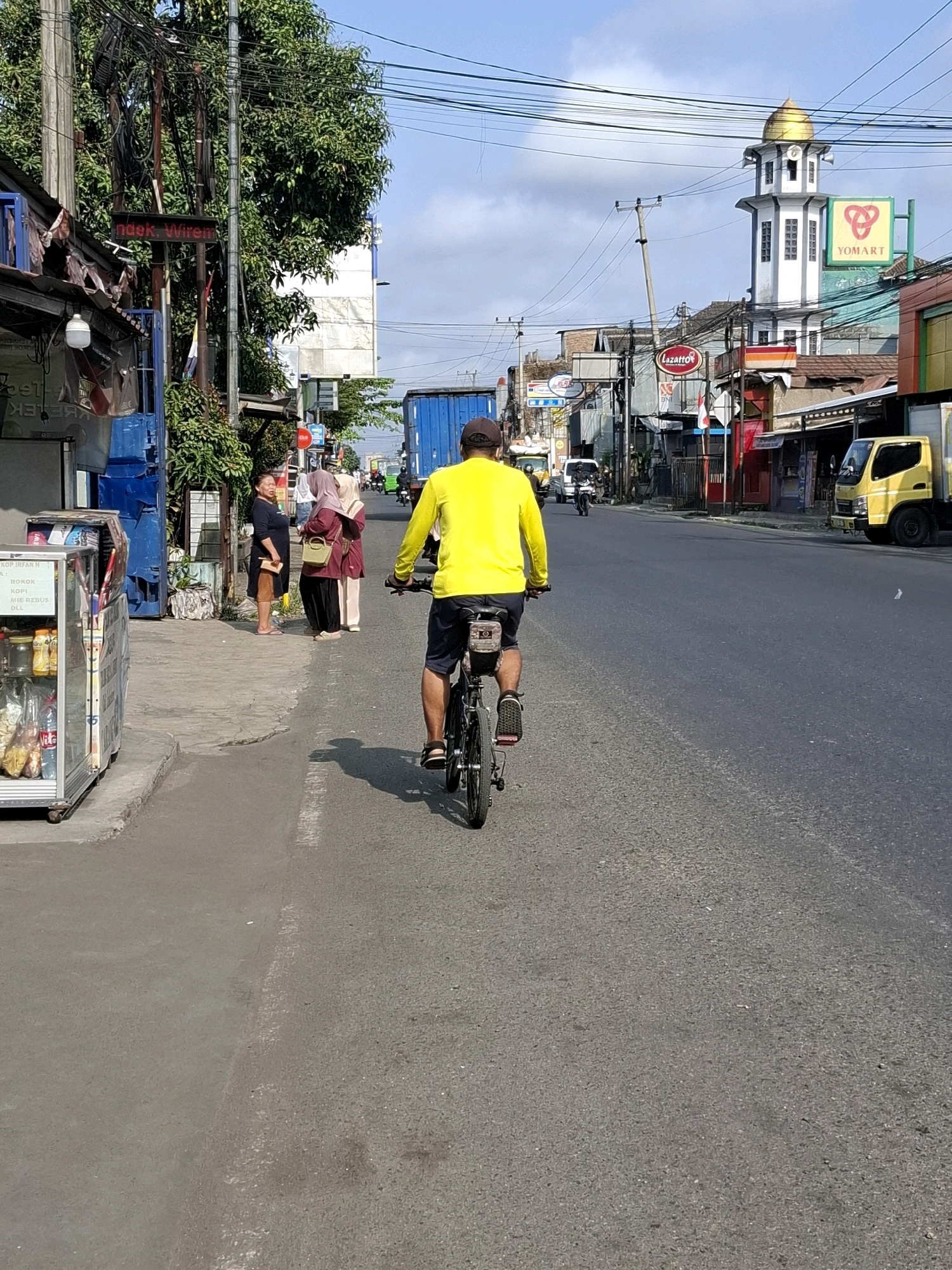 A man wearing a yellow long-sleeved shirt and dark shorts rides a bicycle down a paved road in Sukaraja, Indonesia. He is facing away from the camera, and the bicycle has a black bag attached to its rear. The road is lined with buildings and trees. To the left of the road, there is a sidewalk with a glass-fronted display case filled with goods, and three women are standing on the sidewalk. One woman in a black dress and tan shorts is looking towards the cyclist. Two other women, one in a headscarf and maroon outfit, and another in a light-colored headscarf and pinkish outfit, are standing next to her. Further down the road, a mosque with a golden dome is visible, along with various businesses and a yellow truck. The sky is blue with some clouds.