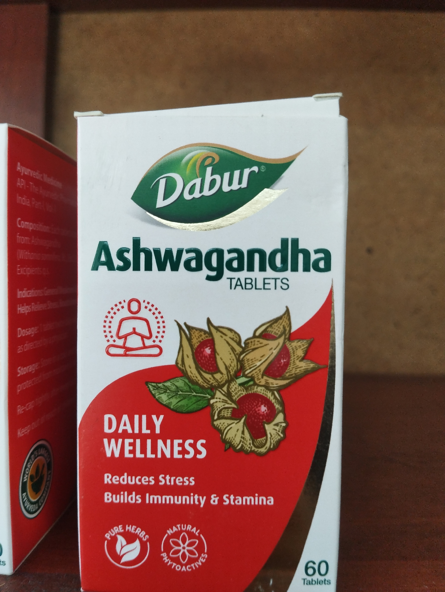 The image shows a close-up of a box of Dabur Ashwagandha Tablets, a dietary supplement. The box is primarily white with red and green accents. The Dabur logo, featuring a green oval with the brand name in white, is prominent at the top. Below the logo, "Ashwagandha TABLETS" is clearly printed. The lower portion of the box displays an illustration of ashwagandha berries and leaves, a red icon of a meditating person, and text that reads "DAILY WELLNESS," "Reduces Stress," and "Builds Immunity & Stamina." There are also circular emblems indicating "PURE HERBS" and "NATURAL PHYTOACTIVES." The number "60 Tablets" is visible at the bottom right. The box is slightly angled and positioned in front of a blurred brown background, possibly a shelf or wall. The image does not contain any information about the city of Tokmok City, Kyrgyzstan, nor does it depict any people or actions.