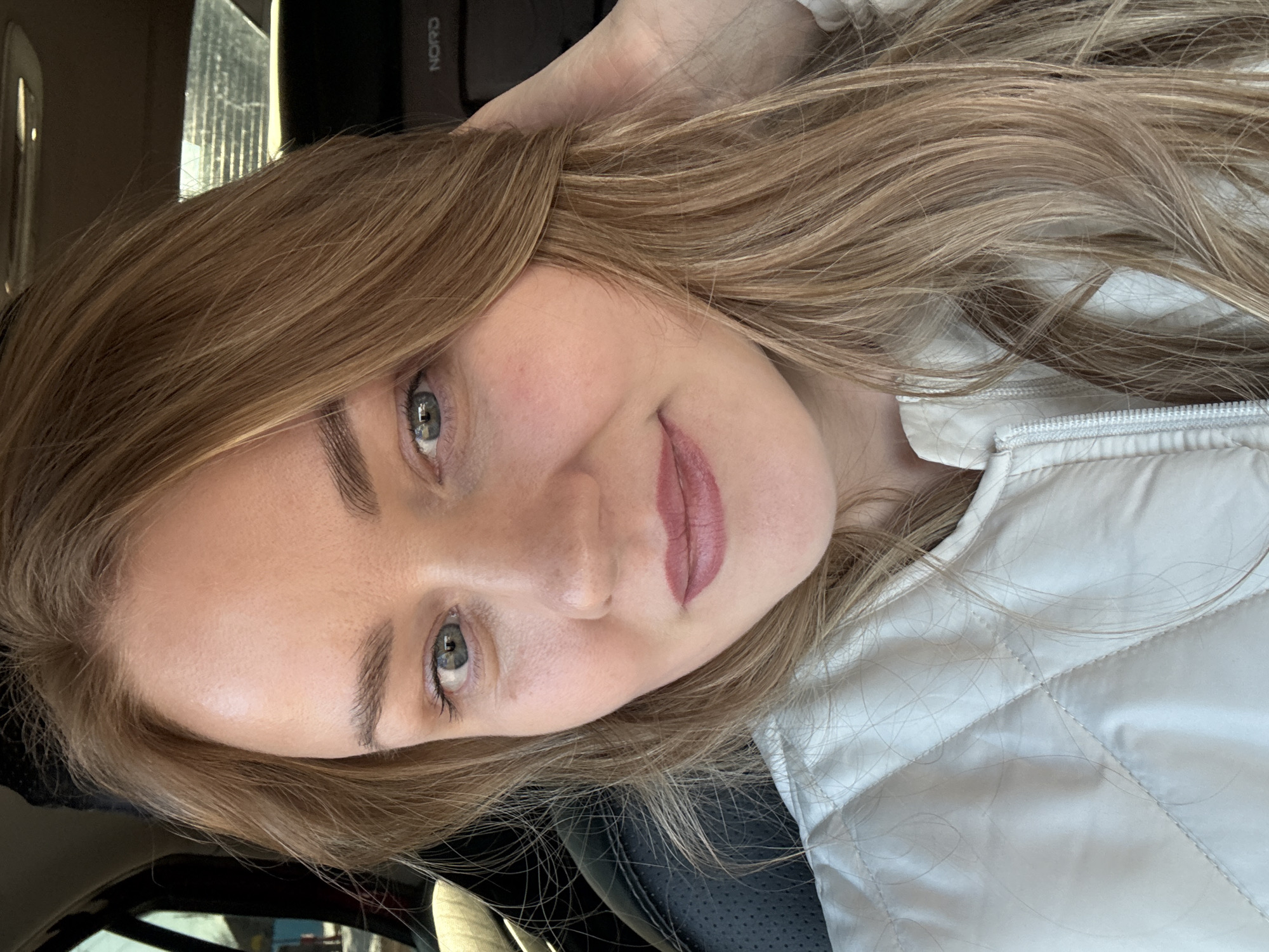 A woman is posing for a close-up selfie, indoors, presumably inside a car. She has fair skin, light brown hair, and light-colored eyes. She is wearing light lipstick and a white quilted vest. The background shows parts of the car interior, including the seat and window. There is no indication of the city Chaikovsky, Russia in the image.