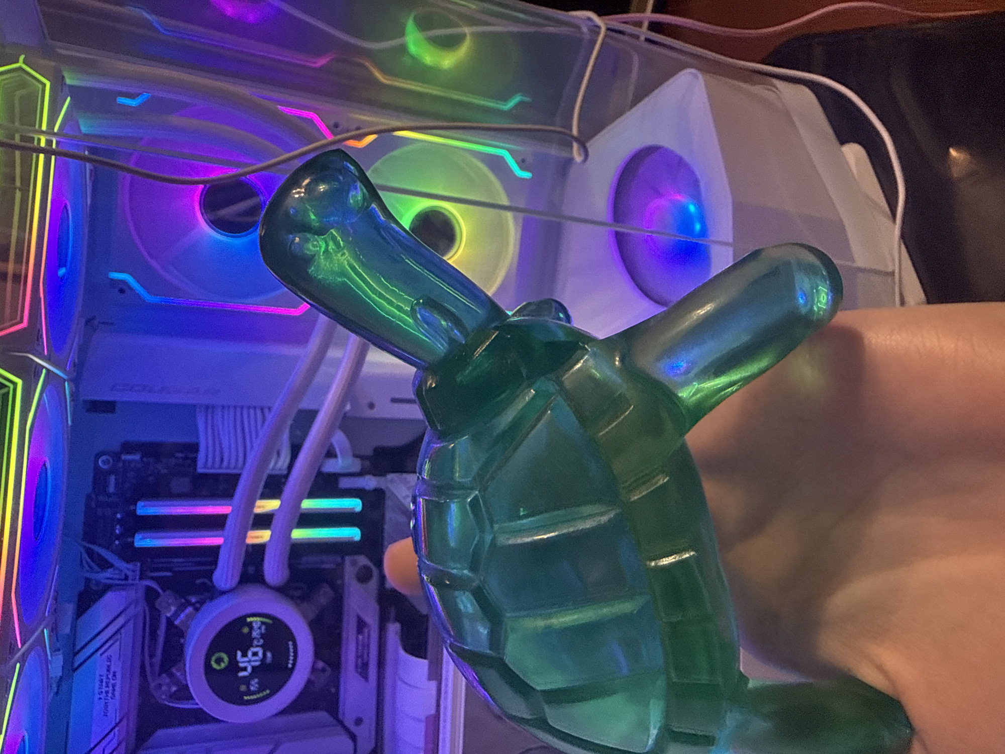 A translucent green turtle figurine is held in a person's hand in front of a computer tower. The computer tower has multiple illuminated fans that display various colors, including purple, pink, and green. The computer's internal components, such as RAM sticks, are also visible and illuminated with colorful LEDs. The background is dark, with some of the computer's internal lighting reflecting off its surfaces and the surrounding environment. The scene appears to be indoors, possibly in a room with controlled lighting.