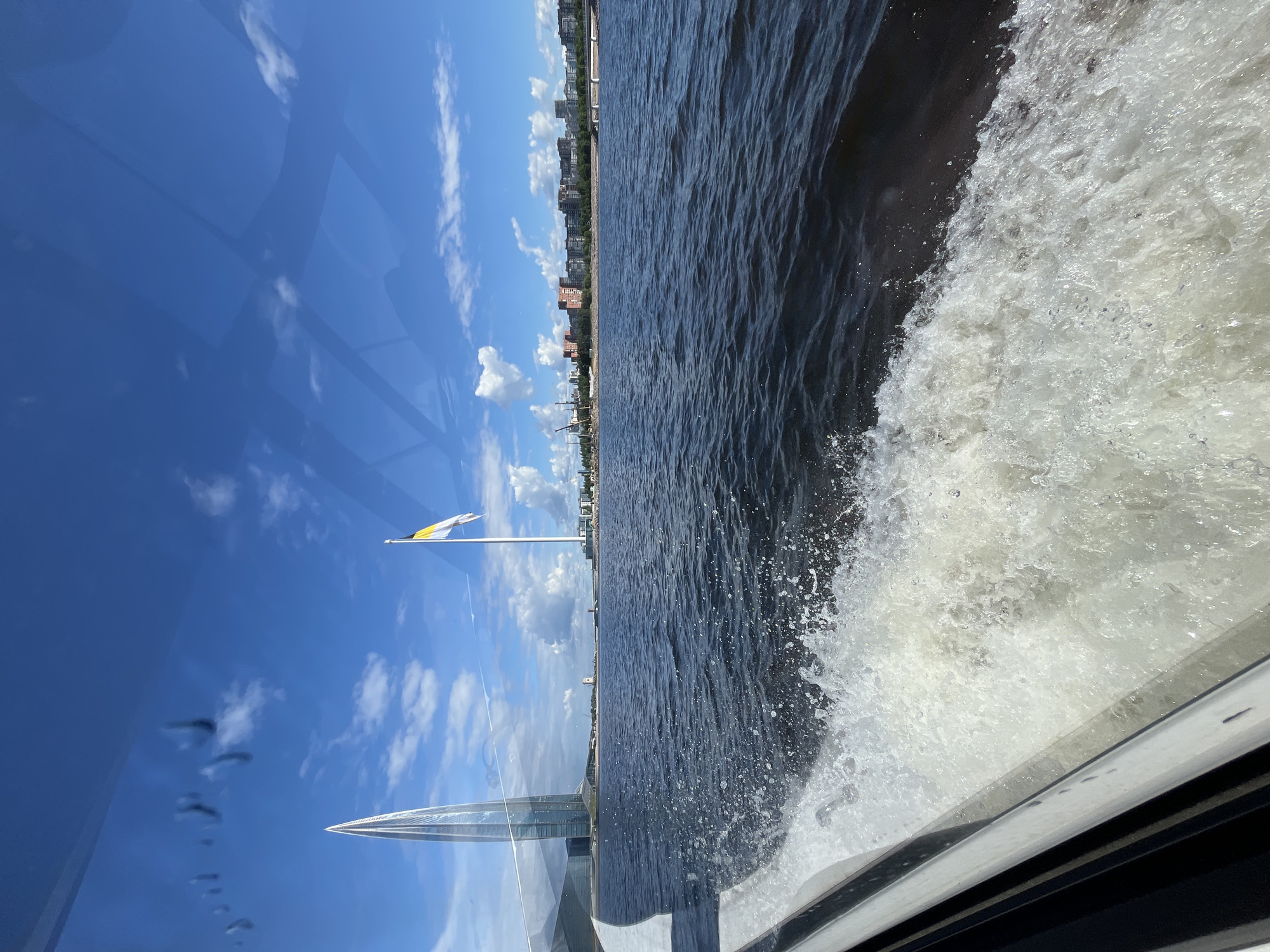 The image shows a view of the Neva River in Saint Petersburg, Russia, from a moving boat. In the distance, the Lakhta Center, a 462-meter-tall skyscraper, dominates the skyline on the left. To its right, a tall flagpole flies a yellow and white flag. The river is dark blue and choppy, with white spray from the boat's wake in the foreground. A row of buildings lines the far bank, and construction cranes are visible in the distance on the left. The sky is a vibrant blue with scattered white clouds. Reflections from water droplets are visible on the glass of the boat's window.
