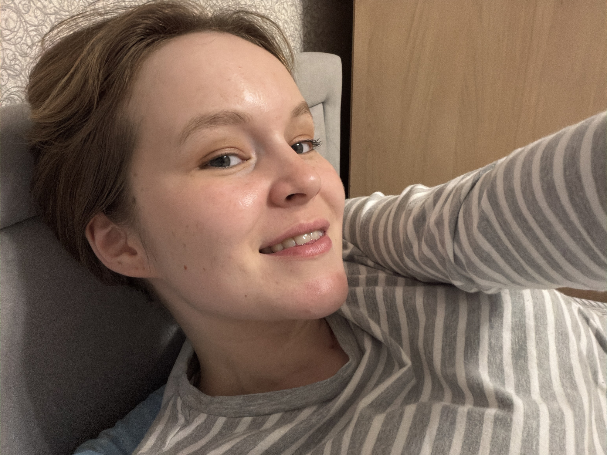 A person with short, light brown hair is lying on a gray surface, possibly a bed or couch. They are wearing a gray and white striped long-sleeved shirt. Their skin appears fair, with some freckles visible. The background includes a light brown surface, possibly a wall or furniture, and a gray headboard. The person is smiling slightly and looking at the camera. There is no verifiable information in the image that confirms the location is Кадряково, Russia. The location is unknown.