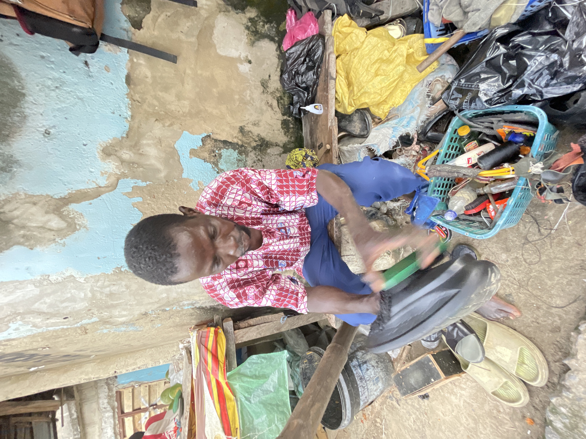 An outdoor scene in Ado, Nigeria, features a man seated on a low wooden bench, engaged in repairing a black shoe. He wears a red and white patterned long-sleeved shirt and blue trousers. In front of him, scattered on the ground, are various items including a pair of tan sandals, a dark-colored shoe, a blue bucket, and a turquoise plastic basket filled with tools and other objects. Behind the man, a wall with peeling blue and beige paint serves as the backdrop, with a wooden structure above it. To the right, a wooden shelf holds black and pink plastic bags, and a small white bottle. Yellow and blue plastic sheeting is also visible. The ground is covered with dirt and debris.