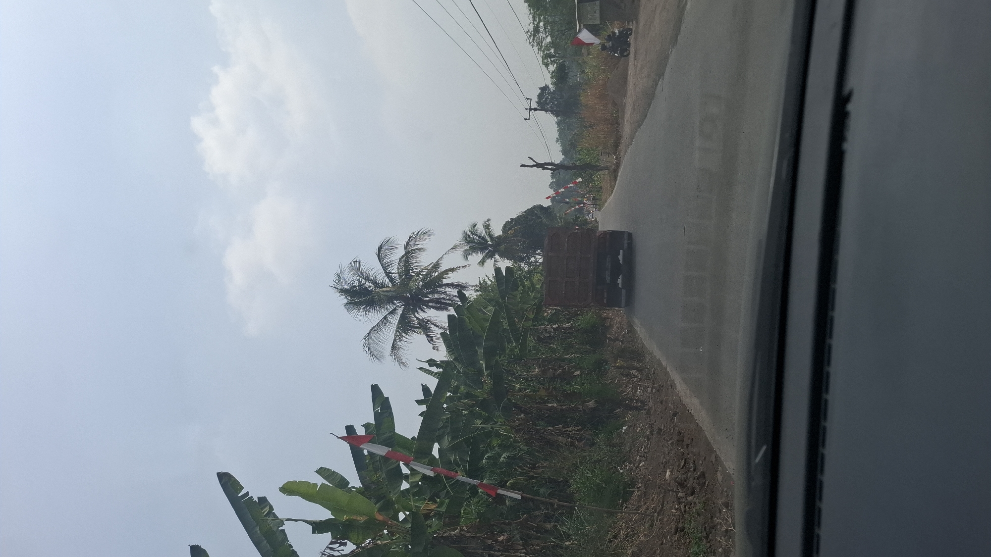 A perspective shot from within a vehicle shows a two-lane paved road in Cibinong Hilir, Indonesia. A brown dump truck is driving away from the viewer down the road, with a few other vehicles and motorcyclists visible in the distance. The sky is overcast with some clouds. The left side of the road is lined with lush green banana trees and palm trees, with several Indonesian flags adorning poles amongst the foliage. The right side of the road features a grassy embankment with power lines and more sparse vegetation. The overall scene depicts a rural highway in Indonesia.