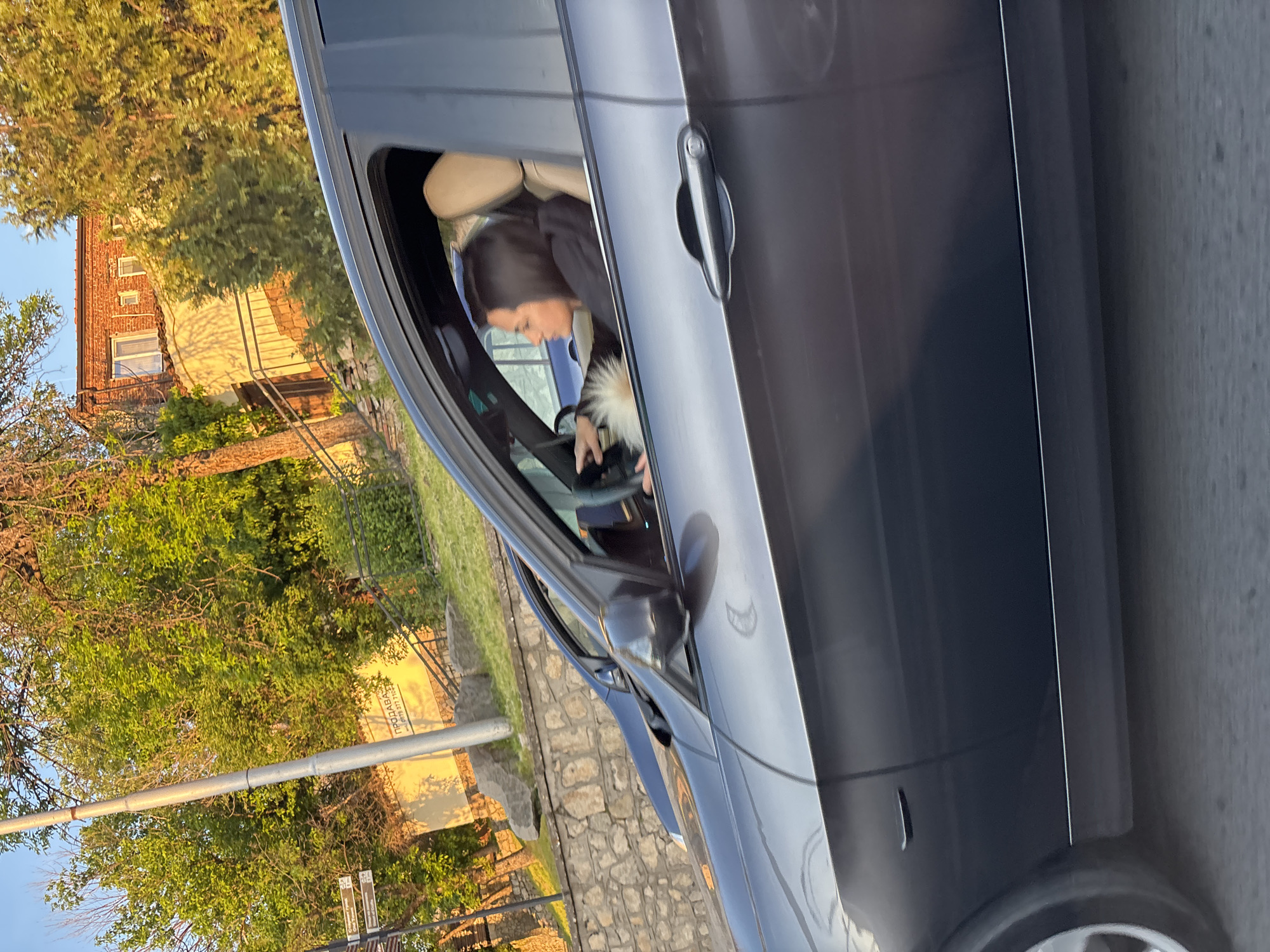 This media file captures a street scene, likely in Plovdiv, Bulgaria, during the late afternoon or early evening, indicated by the warm, golden sunlight and long shadows. The sky is clear blue, suggesting fair weather.
The primary subject is a dark-colored (grey or black) car, likely a sedan, positioned on the right side of the frame. The driver's side front door is partially open or the window is down, revealing a woman with dark hair in the driver's seat. She is wearing a dark top, possibly a jacket, with a prominent fluffy white or light-colored collar or trim. Her head is tilted downwards, and her hands are visible near the steering wheel or dashboard, suggesting she might be operating a device or adjusting something within the car. The car's interior features light-colored (beige or tan) seats.
In the background, on the left side of the image, there is lush green foliage from numerous trees, indicating a warm season. Behind the trees, residential buildings are visible, including a two-story structure with a red-tiled roof and a brick facade, featuring several windows. Another building with a lighter, possibly yellow or stucco, facade is also present, with distinct shadows cast upon it. A stone wall runs along the side of what appears to be a raised property or pathway.
Further back, a grey metal pole stands vertically, and small street signs are visible near its base. Notably, on the yellow wall of one of the background buildings, Cyrillic text is visible, reading "ПРОДАВА" (PRODAVA, meaning "FOR SALE") followed by a phone number "0877 777 764". This text confirms the Bulgarian context provided for the location. The overall setting is an urban or semi-urban residential area.