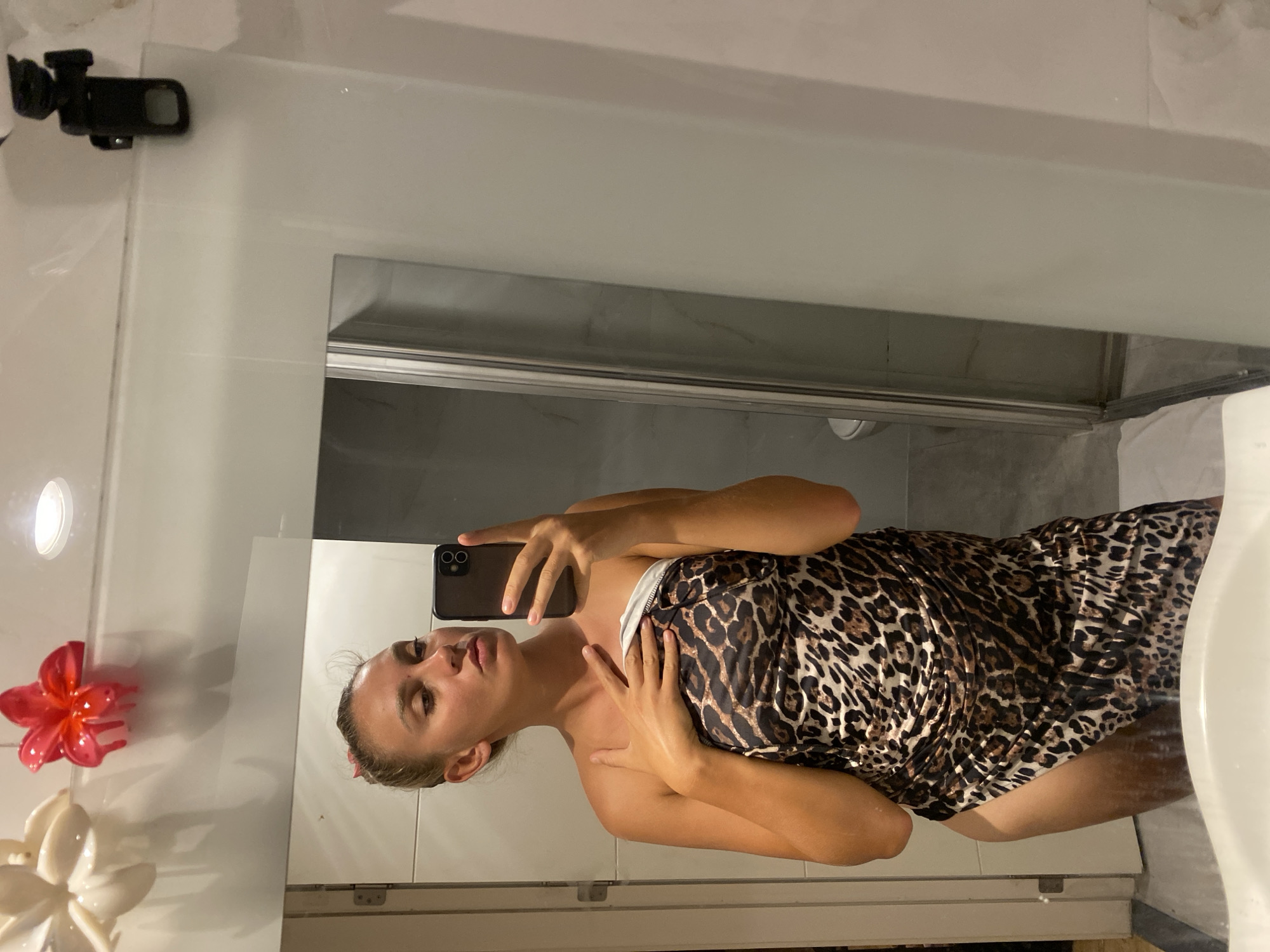 In a bathroom, a young woman takes a mirror selfie. She is wearing a leopard print sarong and has her hair pulled back. She is holding a phone in her right hand, and her left hand is resting on her chest. The bathroom appears to be modern, with white tiles and a glass shower stall. There are also two decorative flower clips attached to the top of the mirror frame. The scene is likely a private residence in Aksu, Turkey.
