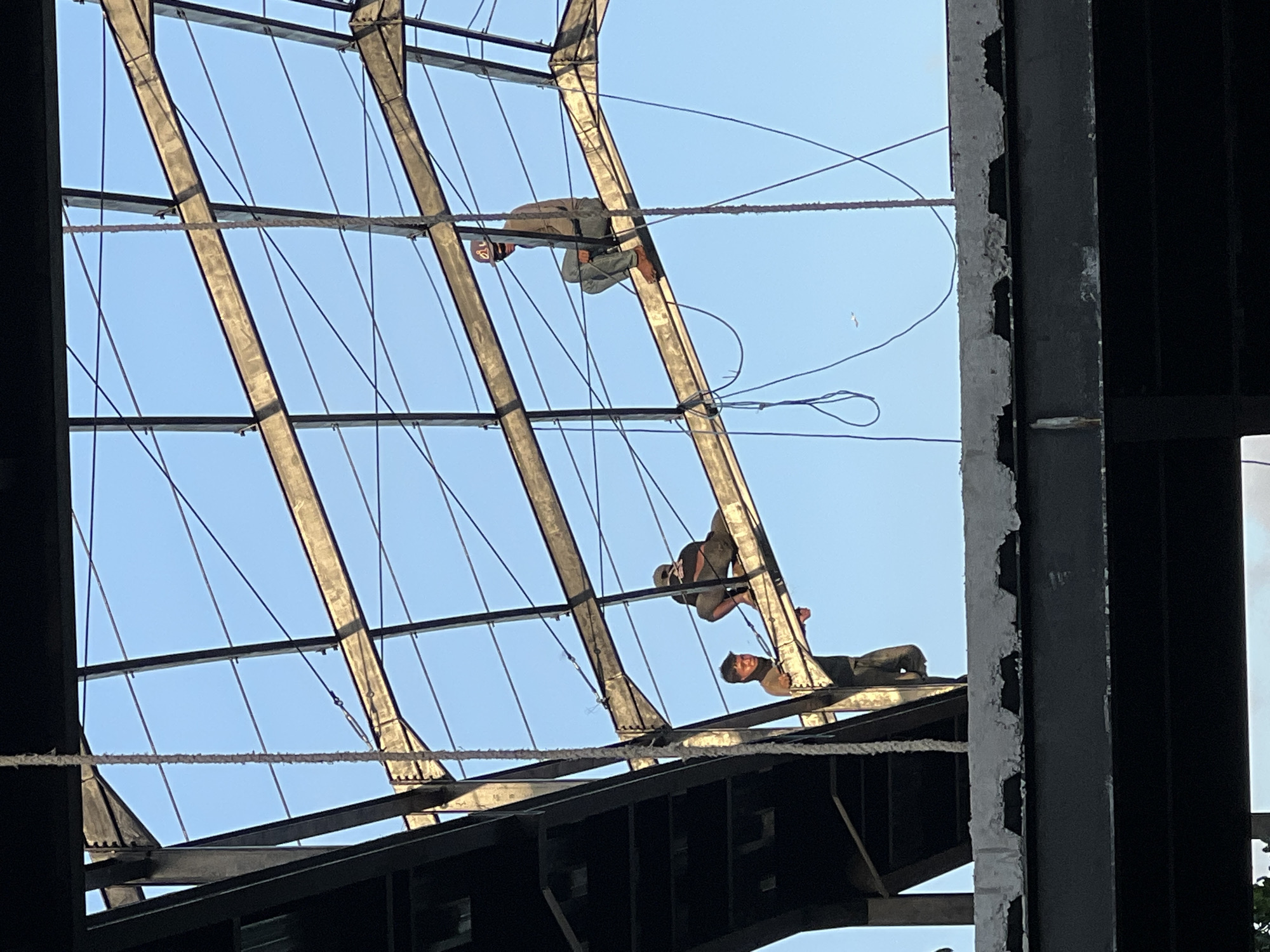 Three workers are perched on the steel framework of a building under construction in Ubud District, Indonesia. The framework consists of a pattern of intersecting beams against a clear blue sky. The workers are secured by safety ropes.