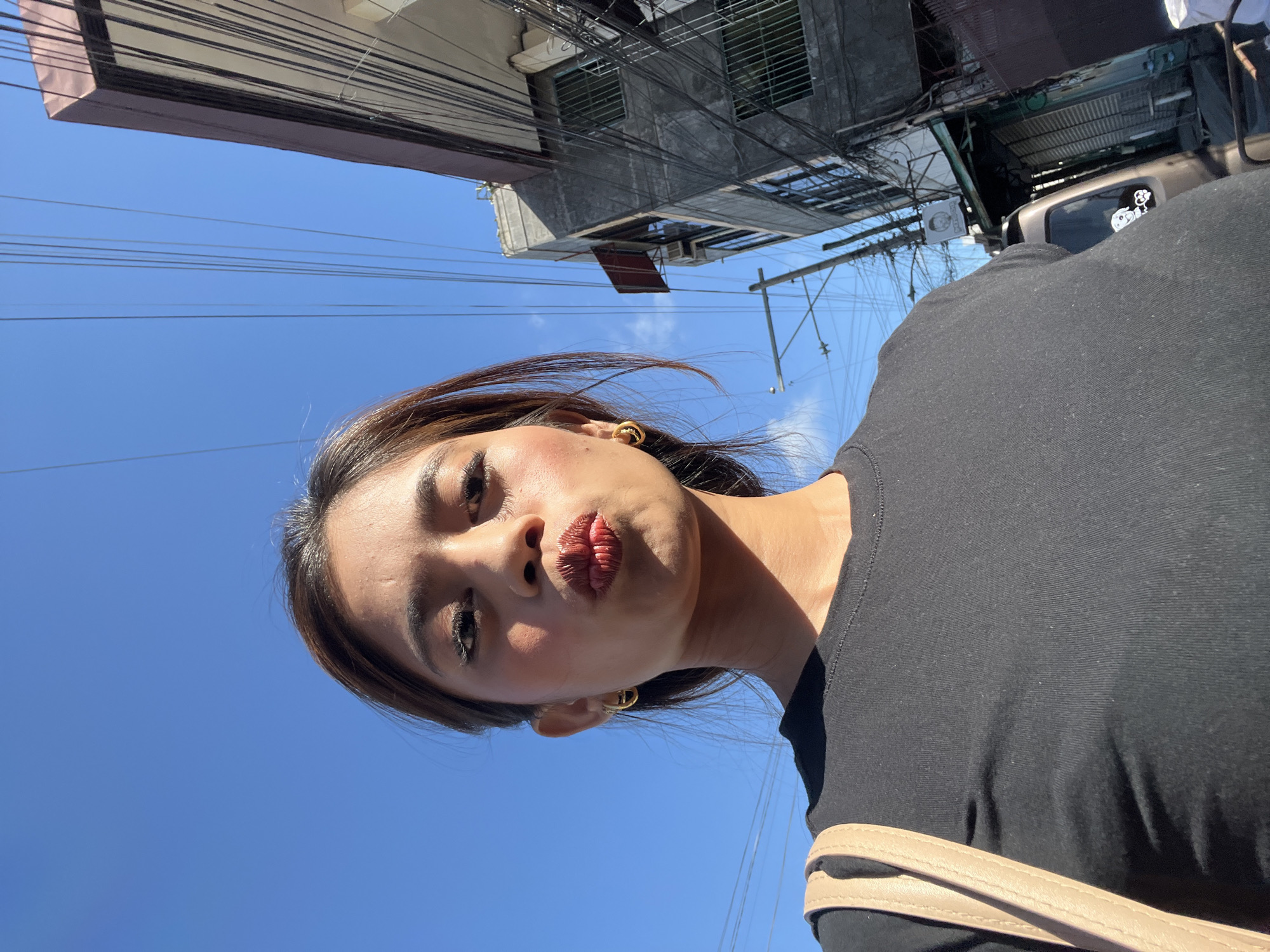 A woman in a black shirt poses for a selfie with her lips pursed, in an outdoor urban setting. The background reveals a clear blue sky with visible overhead electrical wires. To the right, a multi-story building with balconies and numerous wires is visible, suggesting a dense urban environment. The woman is wearing gold hoop earrings and has makeup on, including red lipstick. A light-colored strap from a bag is visible on her left shoulder. The overall scene suggests a sunny day in Davao City, Philippines, characterized by its densely packed buildings and intricate web of electrical infrastructure.