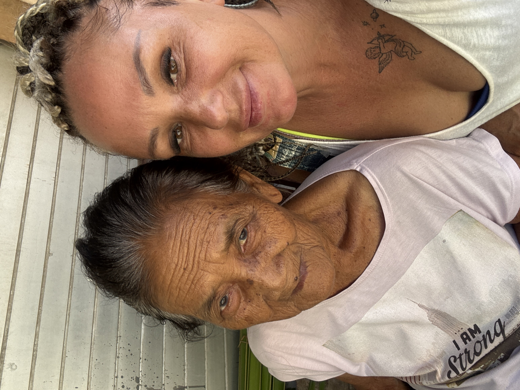 Two women are closely positioned, facing the camera. The woman in the upper position appears younger, with light skin, braided hair, and a tattoo of an angel with a bow and arrow on her upper chest. She wears a white tank top. The woman below appears older, with dark hair and heavily wrinkled skin, wearing a white t-shirt with the words "I AM Strong" on it. They are likely in Kuta, Indonesia, based on the subject. A metallic corrugated wall is visible in the background.