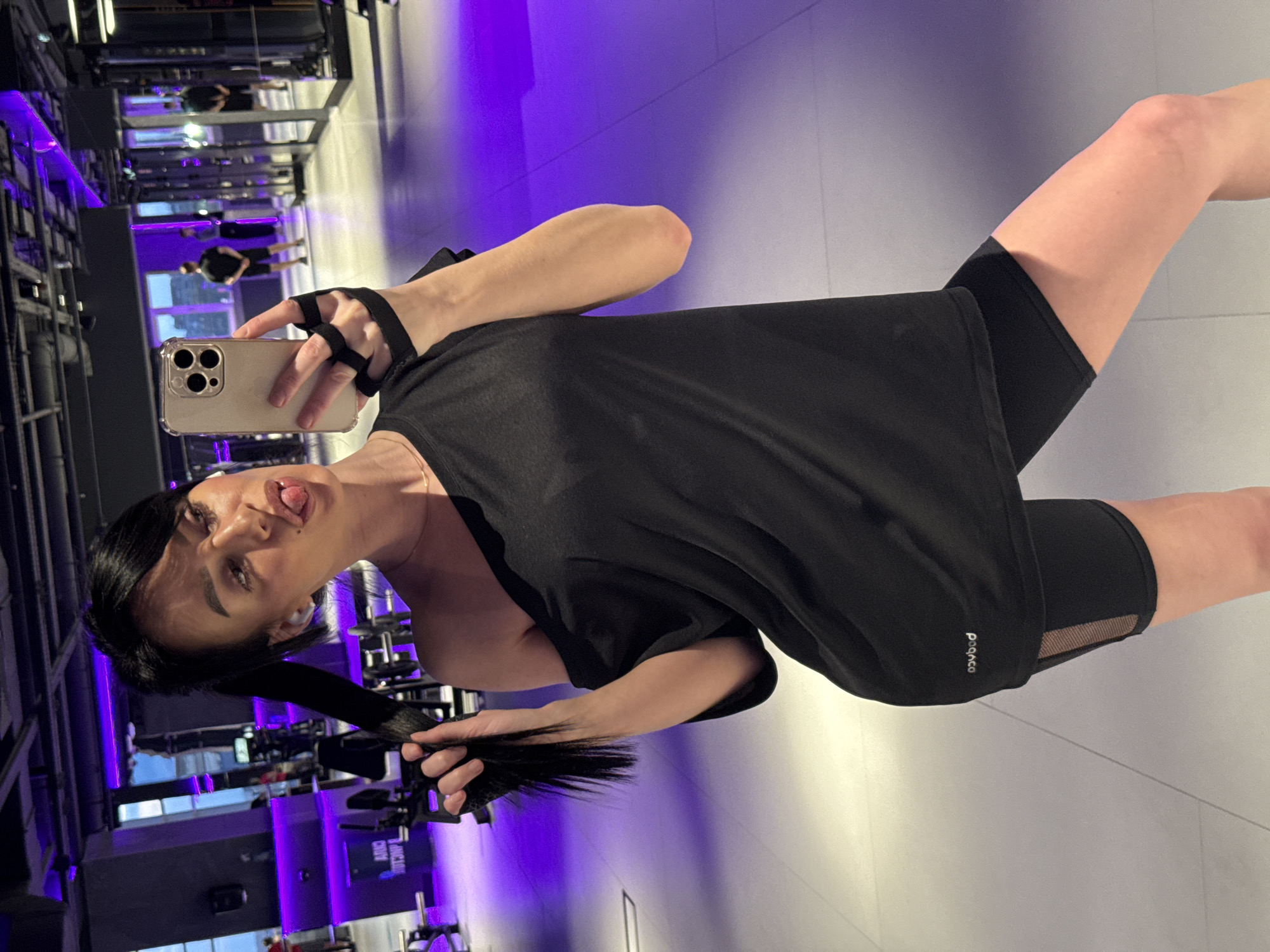 An eye-level selfie shows a young woman posing in what appears to be a modern gym or fitness studio. The image is oriented with the subject's head to the left and feet to the right. She has dark hair, styled in a ponytail that she is holding with her left hand. The woman is wearing a black t-shirt and black cycling shorts, complemented by black fingerless gloves on her right hand, which holds the smartphone used to capture the image. She looks directly at the camera with a playful expression, sticking her tongue out slightly.
The indoor gym setting features dark walls, reflective surfaces, and prominent purple LED lighting that creates a vibrant and contemporary ambiance. Reflections of gym equipment and other individuals are visible in the background mirrors. The floor is light-colored. A white logo, "pogoco", is visible on her black shorts or t-shirt near her right thigh. The artificial lighting inside the gym makes the time of day indistinguishable. The scene depicts a casual moment of a person taking a selfie in a fitness environment, located in Krasnoyarsk, Russia.