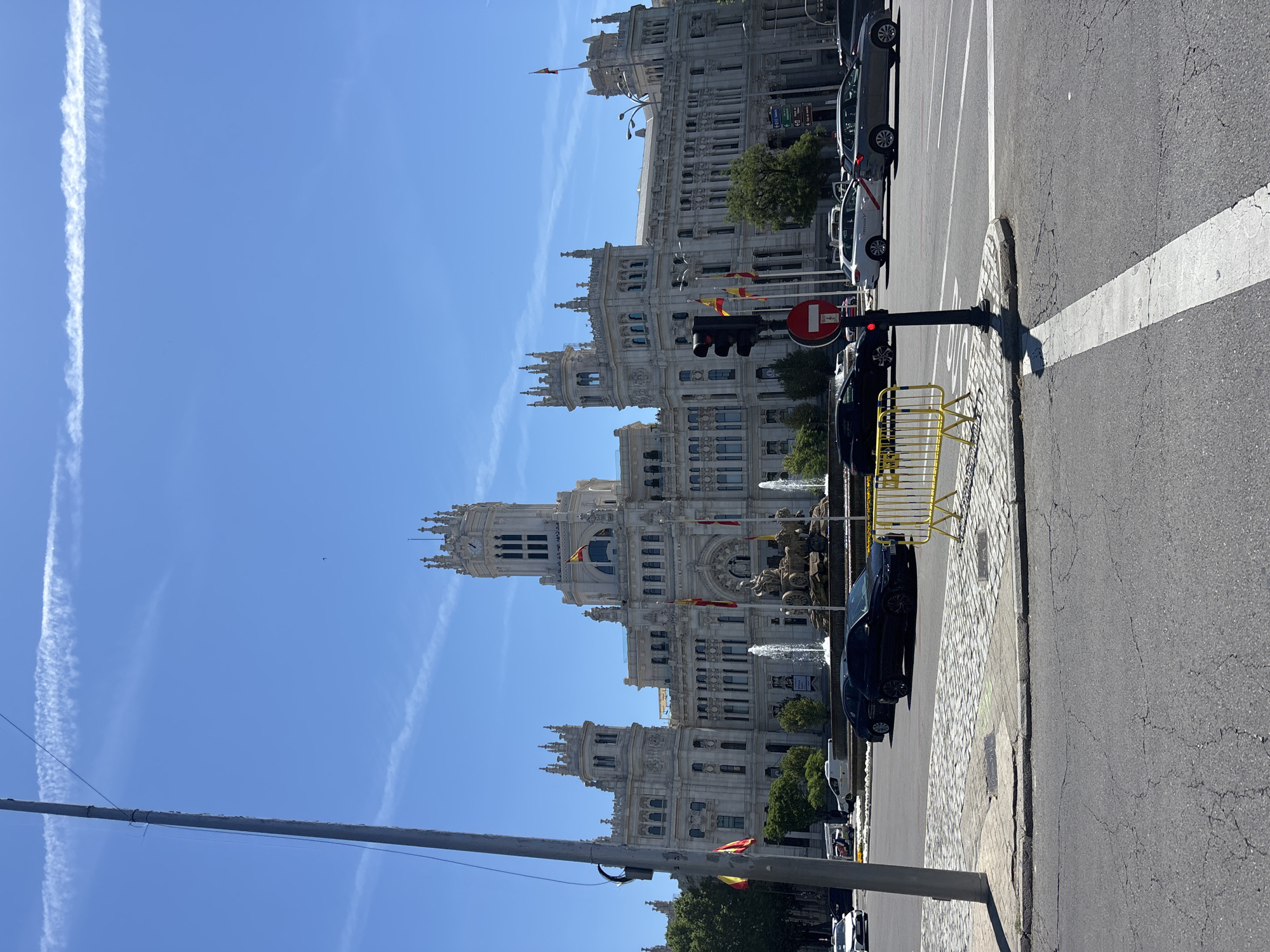 The image shows the Cybele Palace in Madrid, Spain. The building is ornate and light-colored, with a clock tower and multiple spires. Spanish flags are visible on the facade. In the foreground, there is a street with cars parked along the curb. There is a pedestrian crossing with a white line marking it. There is a traffic light with a red light illuminated, and a yellow barrier next to it. The sky is blue with contrails from airplanes. The image is taken from street level.