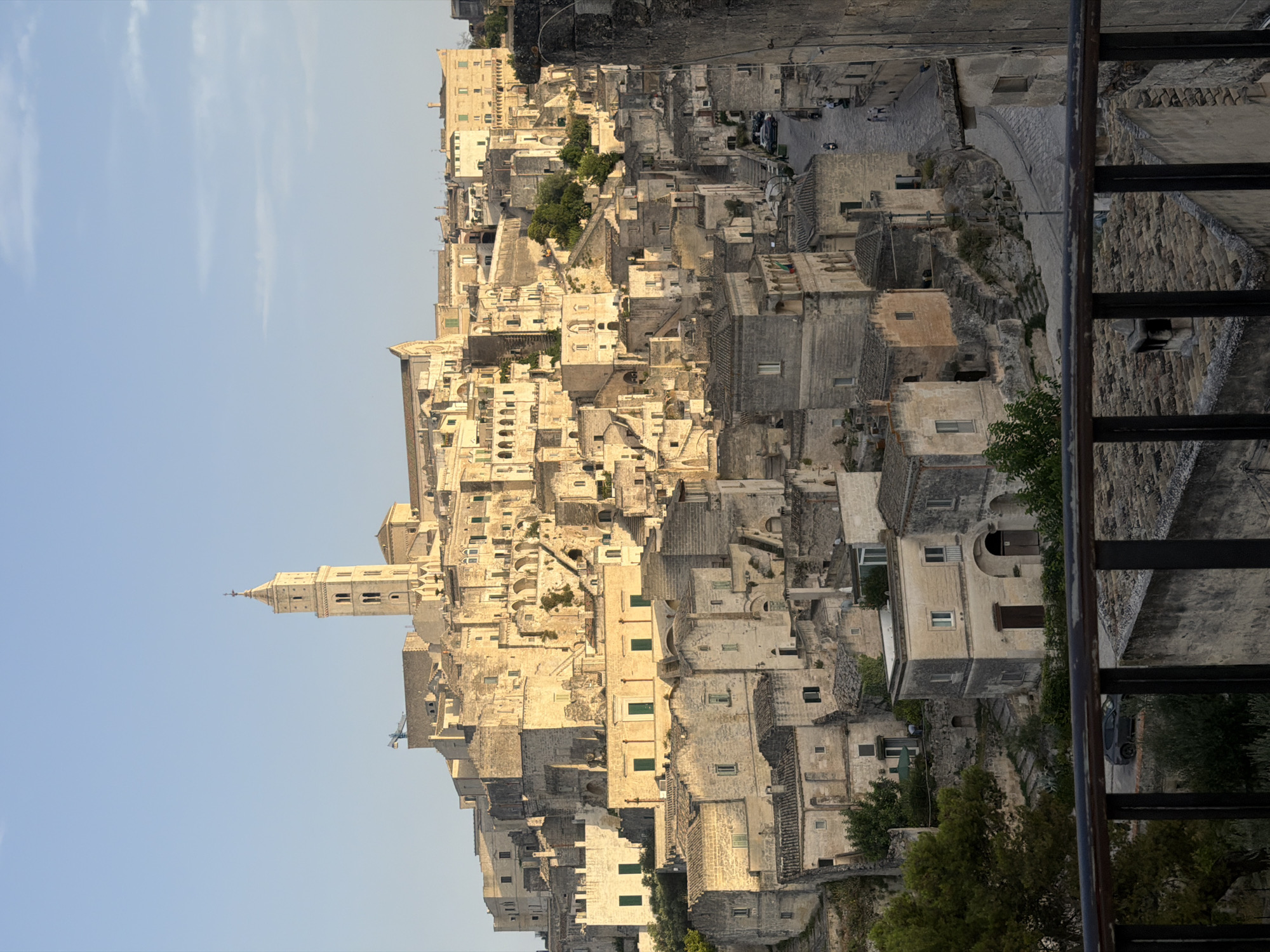 An exterior, eye-level, full shot captures the historic Sassi di Matera in Italy, a UNESCO World Heritage site, bathed in the warm, golden light of a late afternoon sun. The ancient city, carved into the rock, unfolds across a hillside, a dense maze of stone buildings with terracotta roofs. A prominent bell tower, characteristic of the city's architecture, rises above the clustered structures. The sky is a clear, soft blue with a few wispy clouds. In the foreground, a dark metal railing runs horizontally, partially obscuring a view of the lower portions of the city and a stone pathway. The scene is quiet and serene, with no prominent human figures visible, though the presence of parked cars on a winding street and a distant scooter suggest a lived-in, functioning urban environment. The stone buildings exhibit various architectural styles, from simple dwellings to more complex structures with arched windows and doorways, all weathered and aged, bearing testament to centuries of history. The natural landscape is subtly integrated, with some trees and greenery visible amongst the stone, adding a touch of life to the ancient setting.