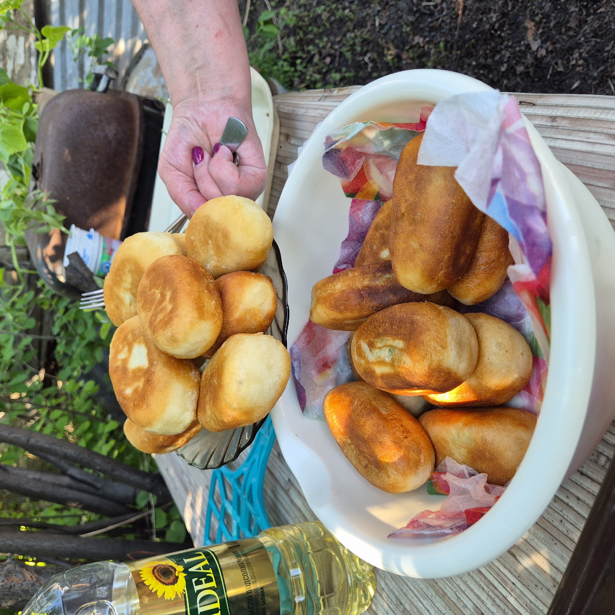 The image depicts an outdoor domestic scene in Новостройка, Russia, captured from a slightly elevated perspective. In the foreground, a large, white, oval-shaped ceramic or plastic bowl holds approximately 15-20 golden-brown, fried pastries, which vary in shape from oval to round. These pastries are resting on crumpled, colorful paper napkins in shades of pink, purple, blue, white, and orange. The bowl is positioned on a light-colored wooden surface composed of parallel planks.
To the left of the bowl, a clear plastic bottle of "IDEAL" brand sunflower oil stands on the same wooden surface. The yellow label features a sunflower graphic and Russian text translating to "Sunflower Oil, Refined Deodorized, Odorless." A bright blue plastic, grid-like object is partially visible between the oil bottle and the large bowl.
Above the large bowl, a person's left hand, adorned with dark pink nail polish, holds a small, clear glass plate with a fluted edge. This plate contains a stack of approximately seven to eight additional golden-brown fried pastries, identical in appearance to those in the large bowl. A silver-colored fork with three tines is visible, either resting among or partially embedded in the pastries on the plate.
The background features dense green foliage, indicating a garden or backyard setting. To the left, dark, gnarled tree trunks are visible. To the right, behind the hand and plate, a large, rusted, dark brown metallic object, possibly an old stove, pot, or heating unit, is discernible. Behind this, a light-colored, corrugated metal fence or wall is present. A small white plastic container is also partially visible near the rusted object. The ground in the bottom right corner appears to be dark soil. Sunlight illuminates the scene, creating highlights on the pastries and the wooden surface.