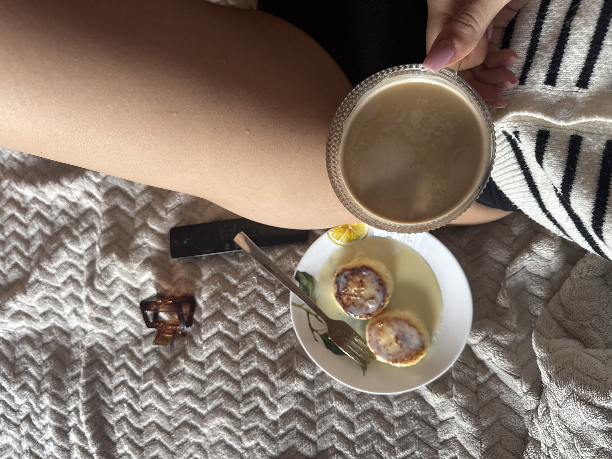 A close-up overhead shot captures a cozy scene on a textured, light-colored blanket. In the lower right, a person's hand with light pink fingernails holds a clear, textured glass cup filled with a light brown beverage, likely coffee with milk. The person is wearing a black and white striped cardigan. To the left of the cup, on a white plate with a subtle floral pattern, are two golden-brown, round pastries drizzled with a white sauce, accompanied by a slice of lemon. A silver fork rests on the plate. In the upper left quadrant of the image, a dark brown hair clip and a black TV remote control are visible on the blanket. The overall lighting suggests an indoor setting, possibly a bedroom or living room, creating a relaxed and comfortable atmosphere, indicative of a breakfast or snack enjoyed at home in Chaikovsky, Russia.