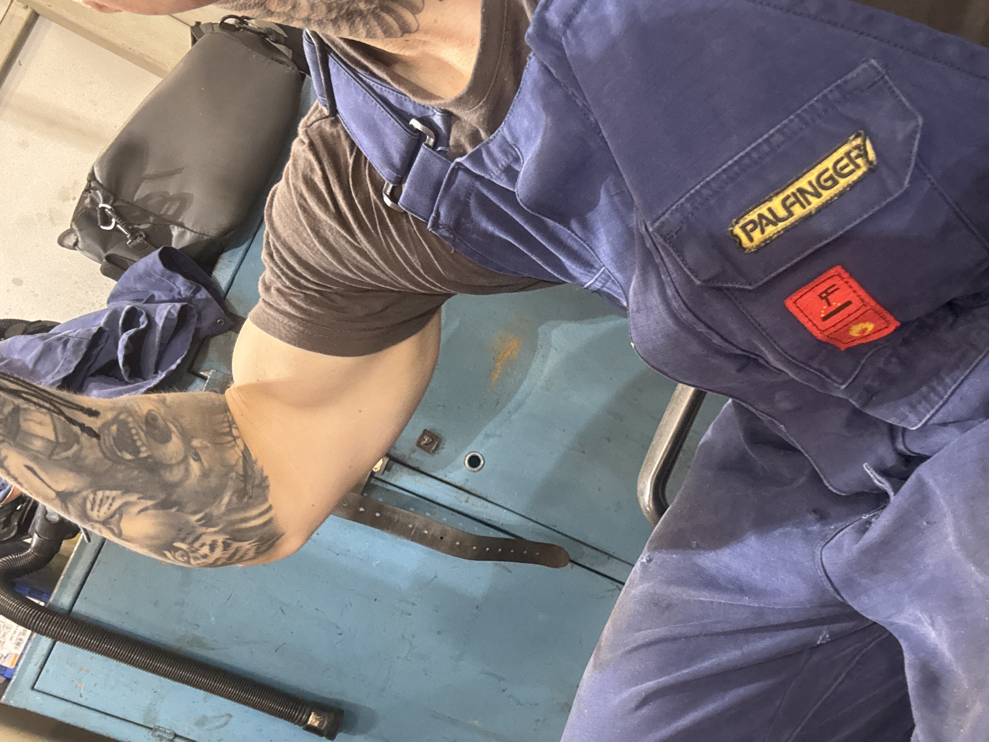 The image shows a close-up of a person's arm and torso, wearing blue overalls and a brown t-shirt. The person has a large tattoo on their left arm. The overalls have a yellow patch with black lettering that reads "PALFINGER" and a smaller red patch with a symbol. The background consists of a blue cabinet and a black bag. The scene appears to be indoors, possibly a workshop or storage area. No specific details regarding the location in Ruptsi, Bulgaria are discernible from the image.
