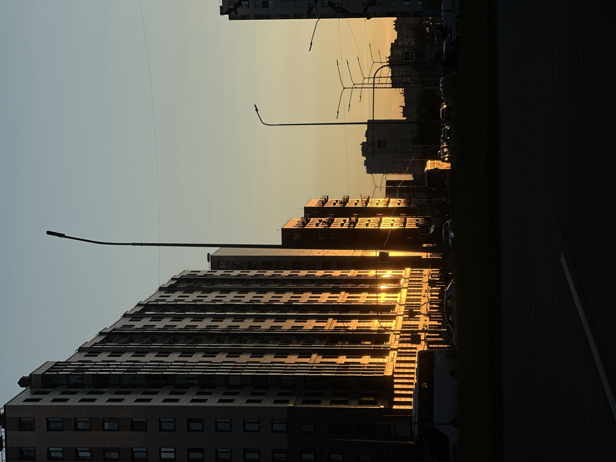 The image depicts a sunset in the city of Otradnoye, Russia. Tall residential buildings with numerous windows are silhouetted against a gradient sky transitioning from pale yellow to light blue. The setting sun casts a warm, golden light on the facades of the buildings on the left side of the frame, illuminating the vertical lines of their architecture.
In the foreground, a dark, grassy median separates a road from the buildings. Several vehicles, including a white bus and various cars, are visible on the road and a sidewalk. Streetlights with modern designs dot the landscape. In the distance, more buildings and infrastructure are visible, further contributing to the urban setting. The overall impression is one of a quiet evening in a residential district of Otradnoye, with the striking effect of the sunset enhancing the cityscape.