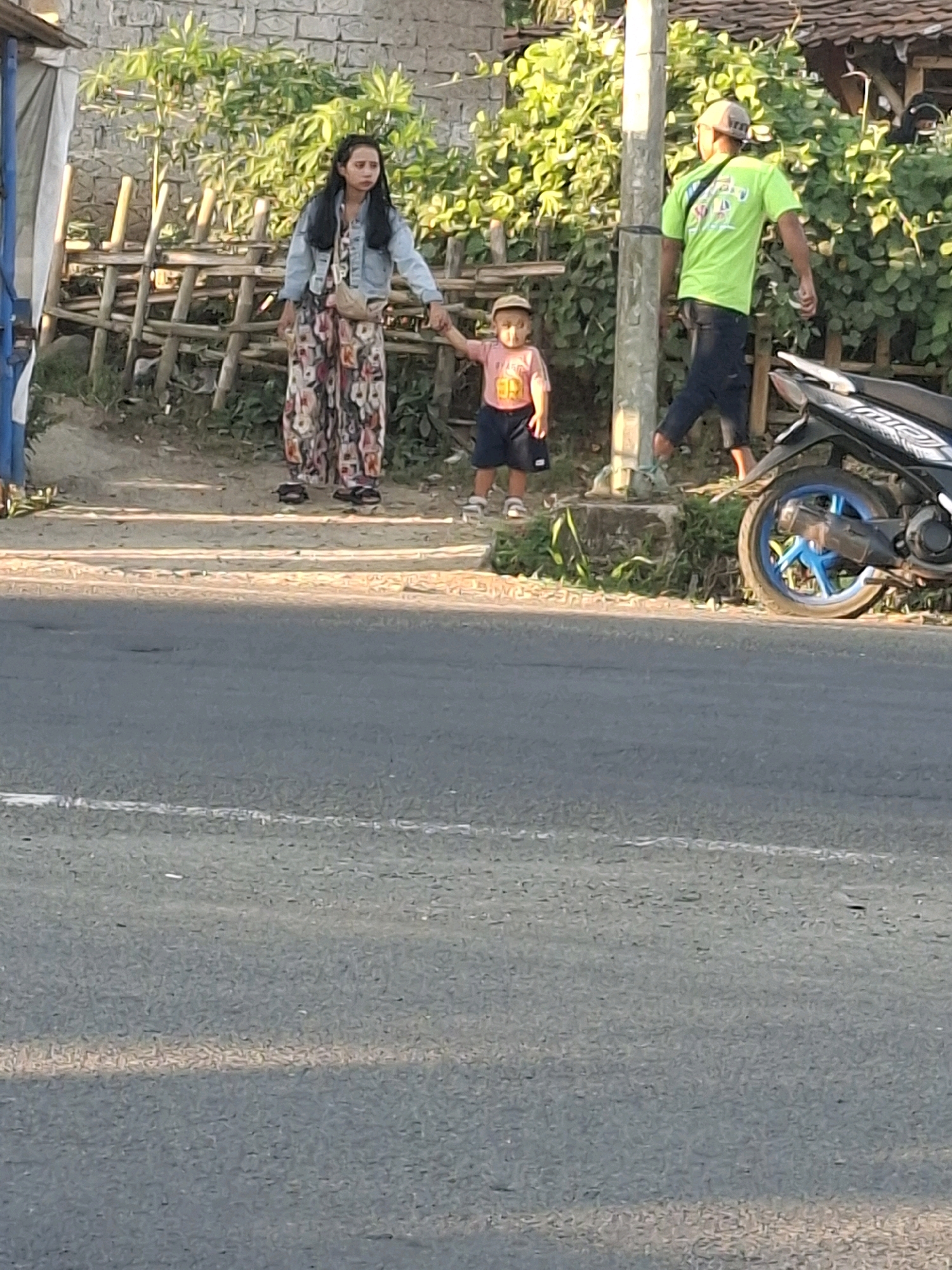 In Cikaret, Indonesia, on a paved road bordering a dirt path, a woman in a long floral dress and a denim jacket holds the hand of a young child wearing a pink shirt and dark shorts. The child is looking to the side with their mouth open. Behind them, a man in a bright green t-shirt and dark shorts walks away from the camera. A parked motorcycle with a blue-rimmed wheel is visible on the right side of the frame. The background features a stone wall, a wooden fence, and lush green foliage. The scene is illuminated by natural sunlight.