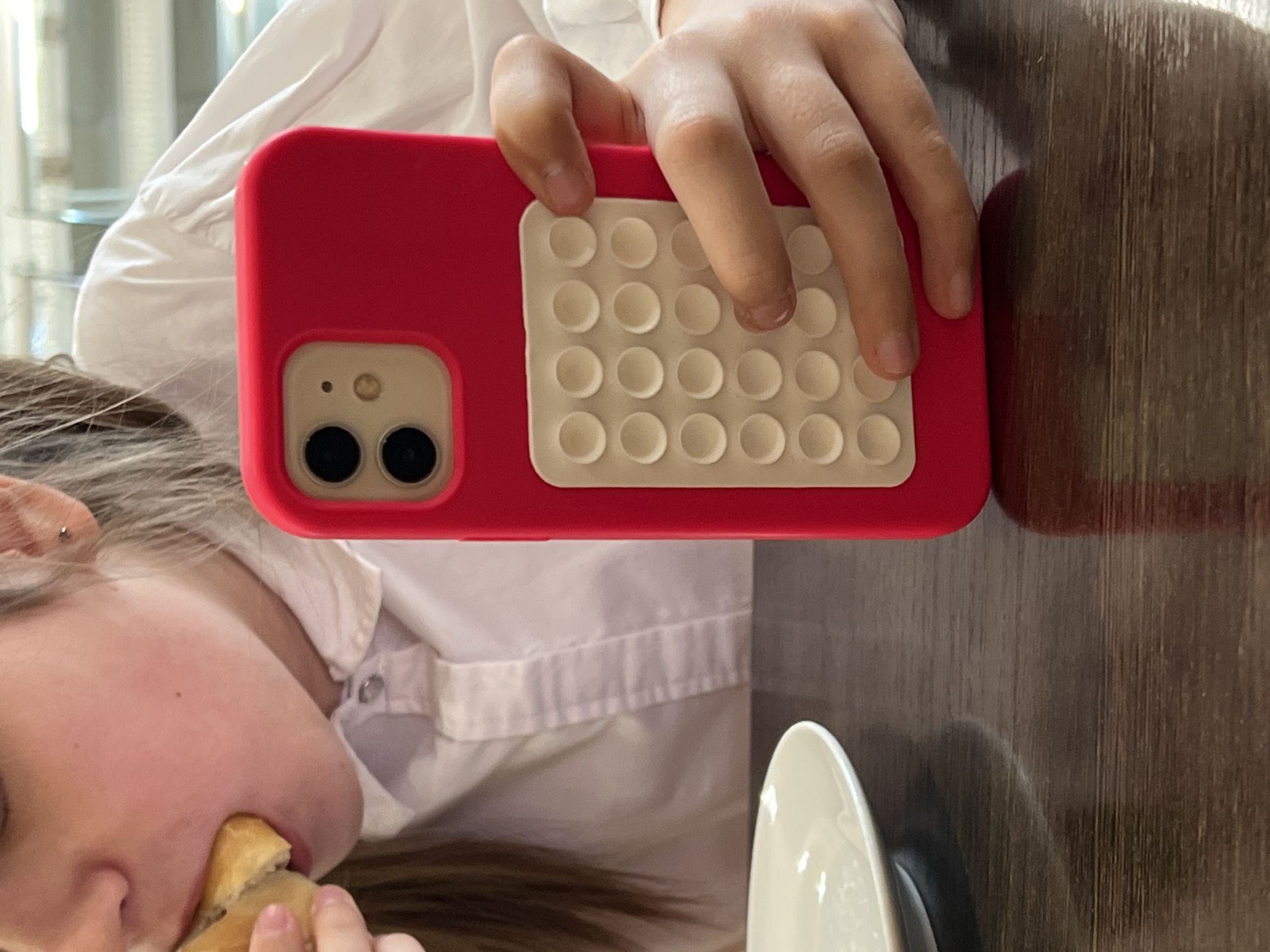 A person is eating while holding a red iPhone with a white pop fidget toy on the back. The person is wearing a white button-down shirt. A white plate is on a dark wooden table. The location is not discernible from the image and cannot be identified as Chaikovsky, Russia.