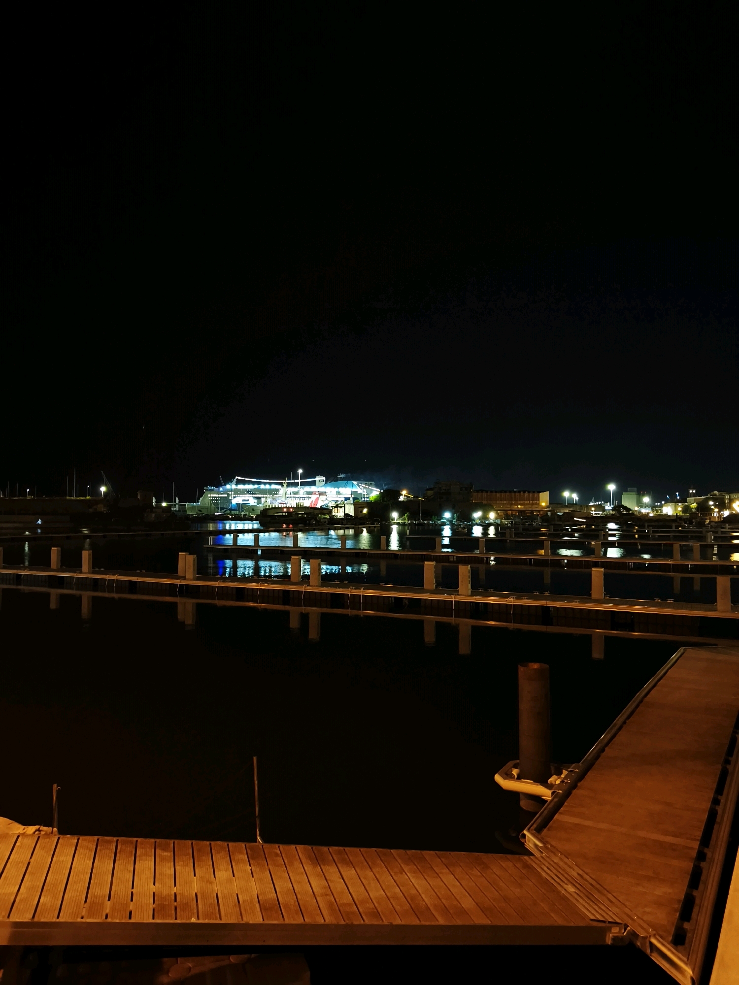 This is a long shot, nighttime photograph taken at the port of Livorno, Italy. In the background, a large illuminated ferry or cruise ship is docked, its lights reflecting on the dark water. The foreground features a wooden pier, with several other piers extending into the water. The scene is dark with artificial lighting from the ship and distant buildings, creating reflections on the water. There are no people or visible animals in the image.