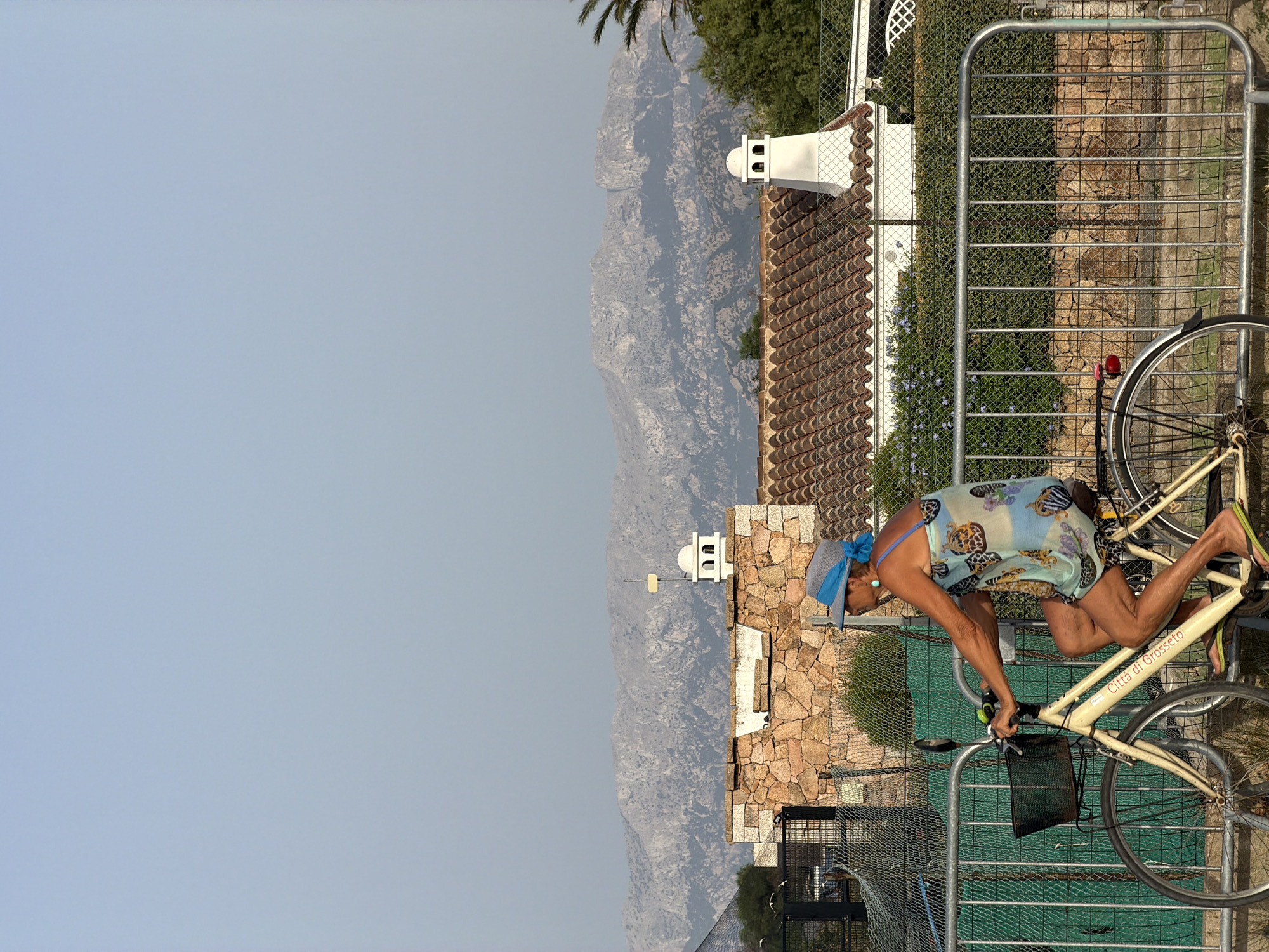 An elderly woman wearing a blue headscarf and a light blue, patterned dress rides a cream-colored bicycle. The bicycle has a basket on the front and is labeled "Città di Grosseto". She is wearing flip-flops. The woman is in profile, facing right, and appears to be moving. She is riding on a paved surface next to a metal fence. Behind the fence, there is a stone wall and a building with a terracotta tiled roof. In the background, there are mountains under a hazy sky. The scene suggests a location in or near a Mediterranean coastal town, possibly Olbia, Italy, given the style of the architecture and the presence of mountains and a pale sky.