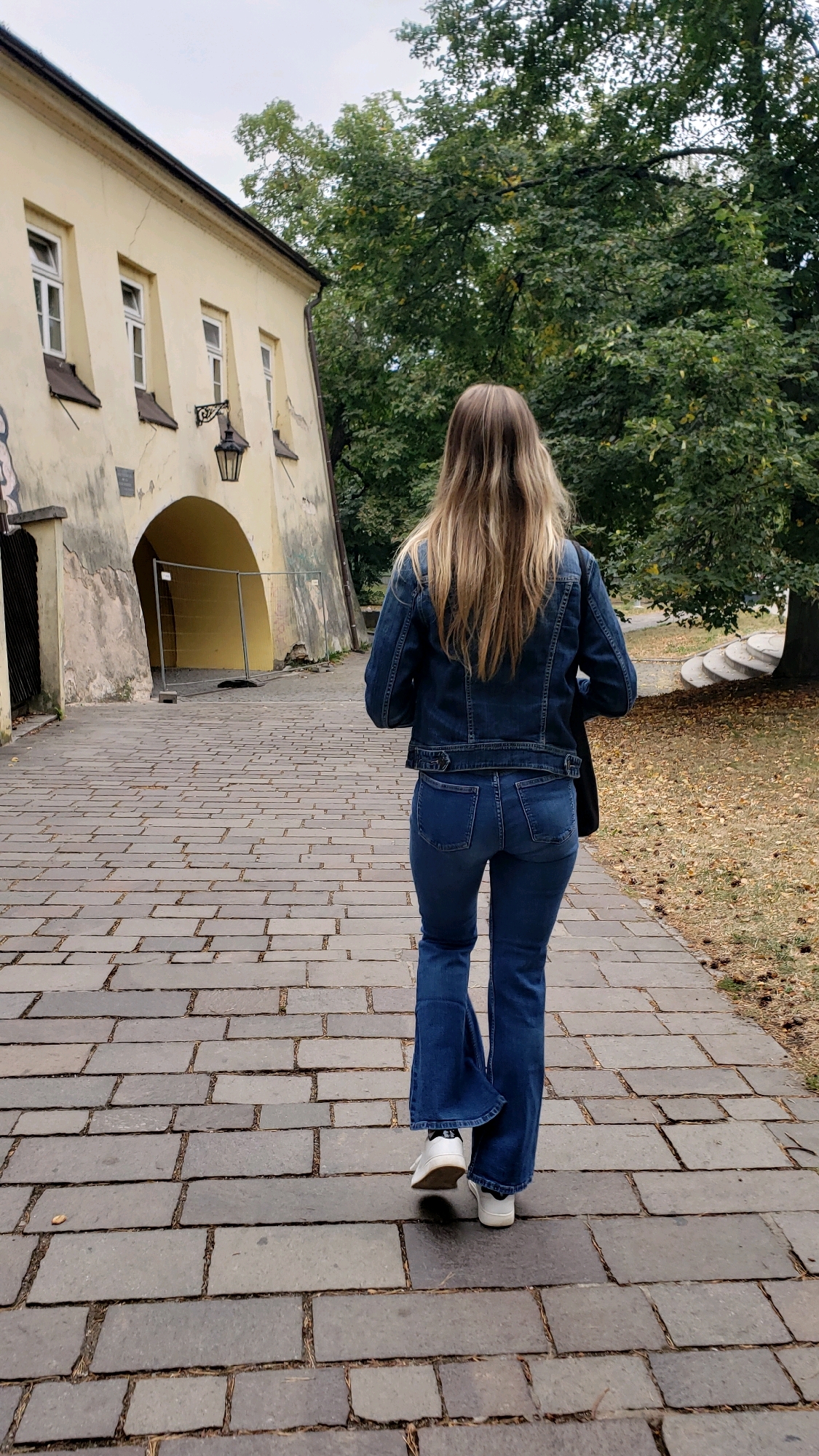 The image shows a woman from behind, walking on a cobblestone path towards a large, arched gateway. She is wearing a blue denim jacket, blue flared jeans, and white sneakers. Her blonde hair is long and extends down her back. To the left of the path is a light yellow building with a dark arched opening, behind which a metal barrier is visible. A street lamp is mounted on the building above the arch. To the right of the path, there are trees with green and yellowing leaves, and fallen leaves on the ground, suggesting an autumn setting. The path itself is made of large, rectangular grey cobblestones. In the distance, on the right side of the image, there are stone steps leading up. The overall setting appears to be a historical or park-like area within the city of Prešov, Slovakia.