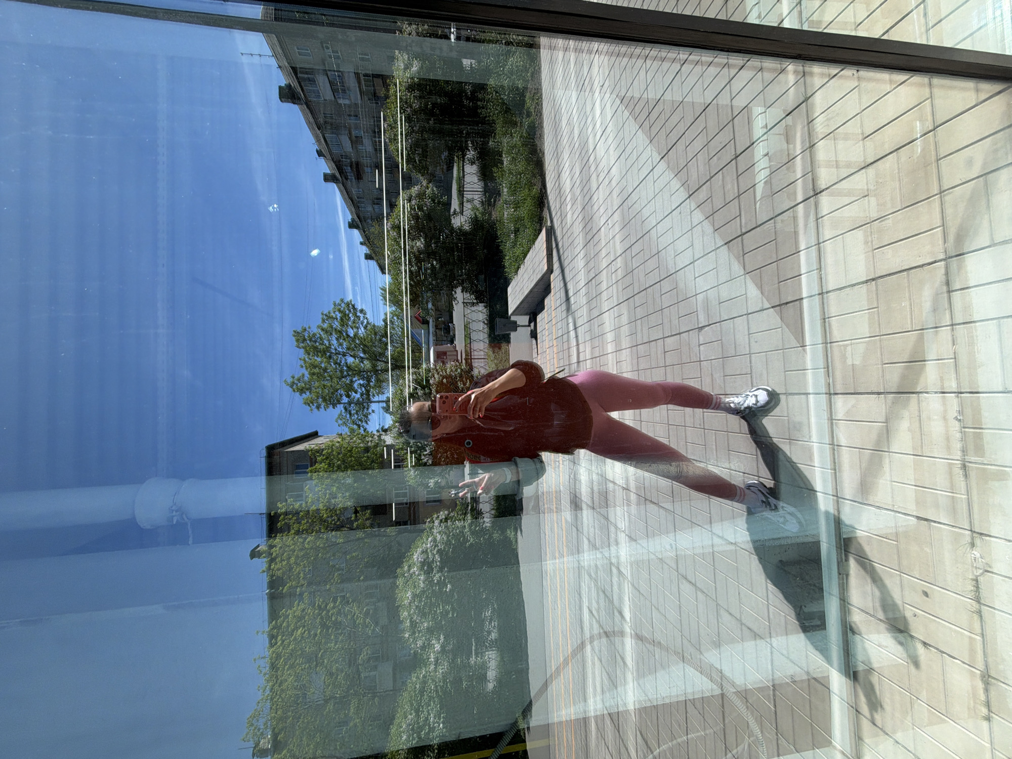 The image shows a reflection of a person taking a photograph in a window. The person is wearing a dark shirt, pink leggings, and white sneakers. They are holding a pink phone. The reflection in the window also shows a building, trees, a blue sky, and a paved area. The location is Birobidzhan, Russia.