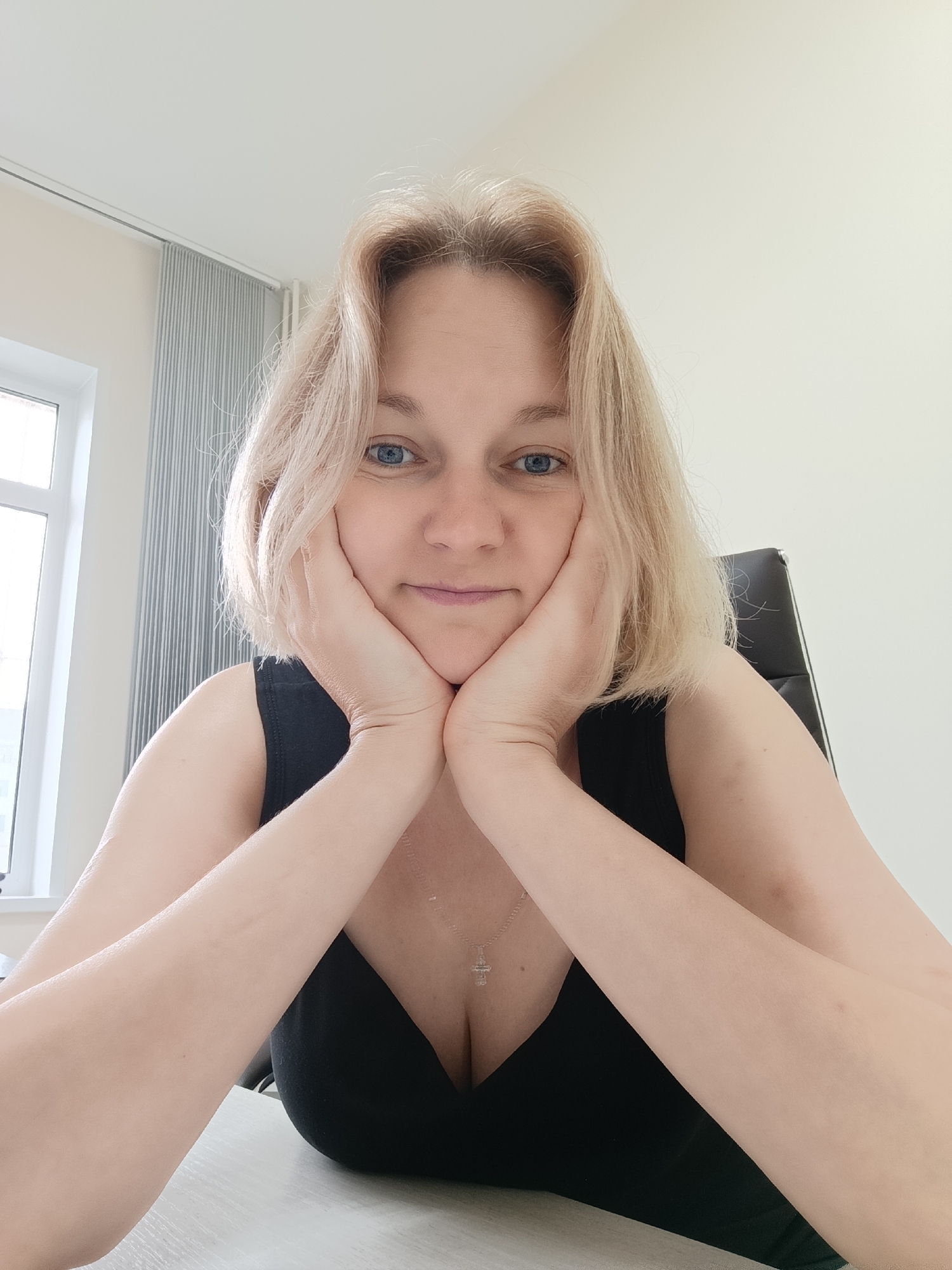 In a well-lit room, a Caucasian woman with blonde hair and blue eyes is seated at a desk, her hands resting on her cheeks. She is wearing a black tank top and a silver necklace with a cross pendant. A black office chair is visible behind her. To the left, a window with vertical blinds is present, and the room has light-colored walls and ceiling. The overall impression is that of an indoor setting, possibly an office or a home study, with no elements to suggest a specific location in Markovsky, Russia.