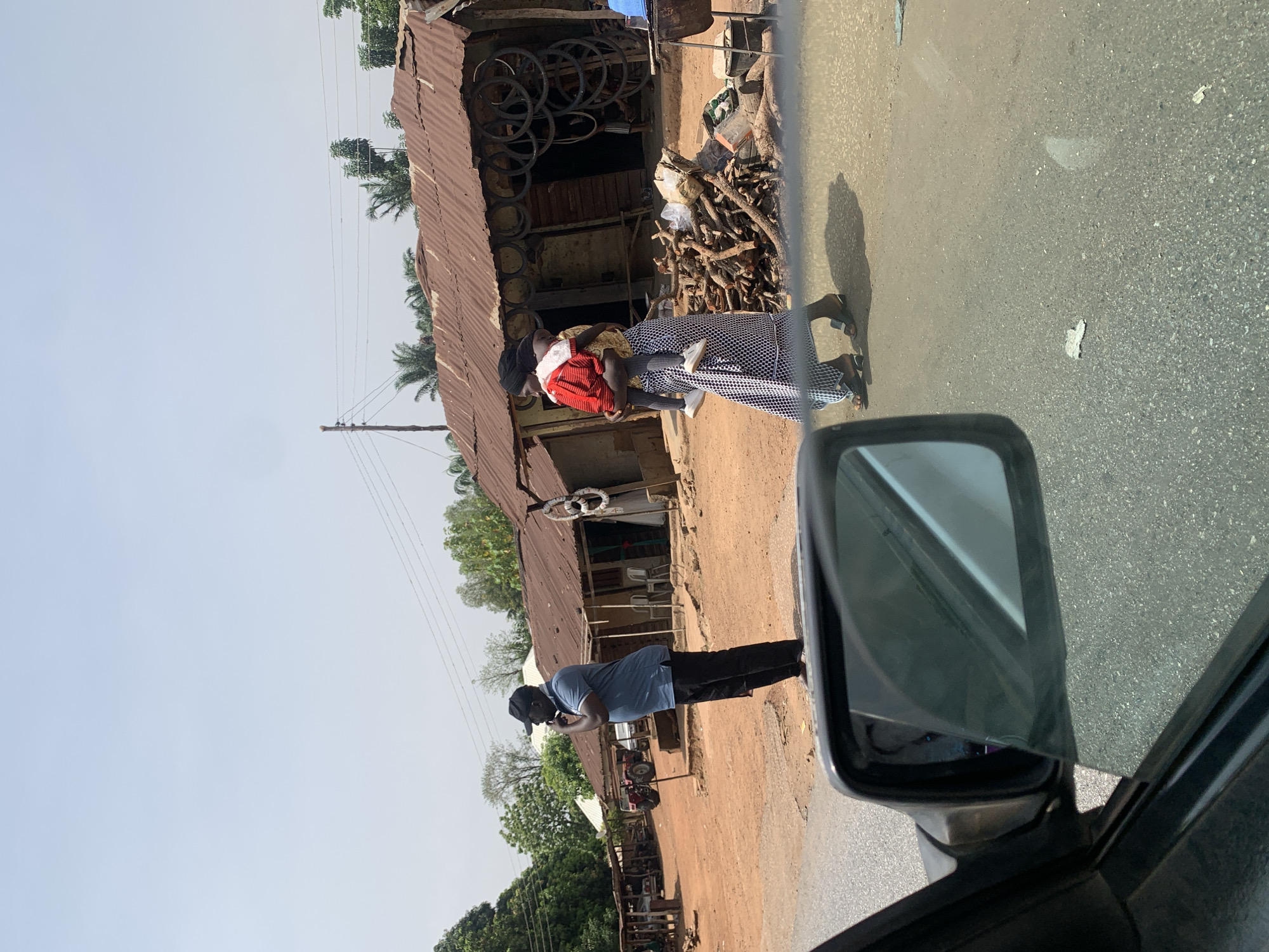 The image depicts a street scene in Karshi, Nigeria. A woman wearing a red shirt and carrying a child stands in front of a corrugated metal-roofed building, possibly a shop. The building has tires and firewood stacked near its entrance. A man in a blue shirt and black pants stands nearby, appearing to be using a phone. The scene is viewed from inside a vehicle, with the side mirror visible in the foreground. The street is paved and adjacent to a dirt area with other structures and vegetation visible in the background under a clear sky.