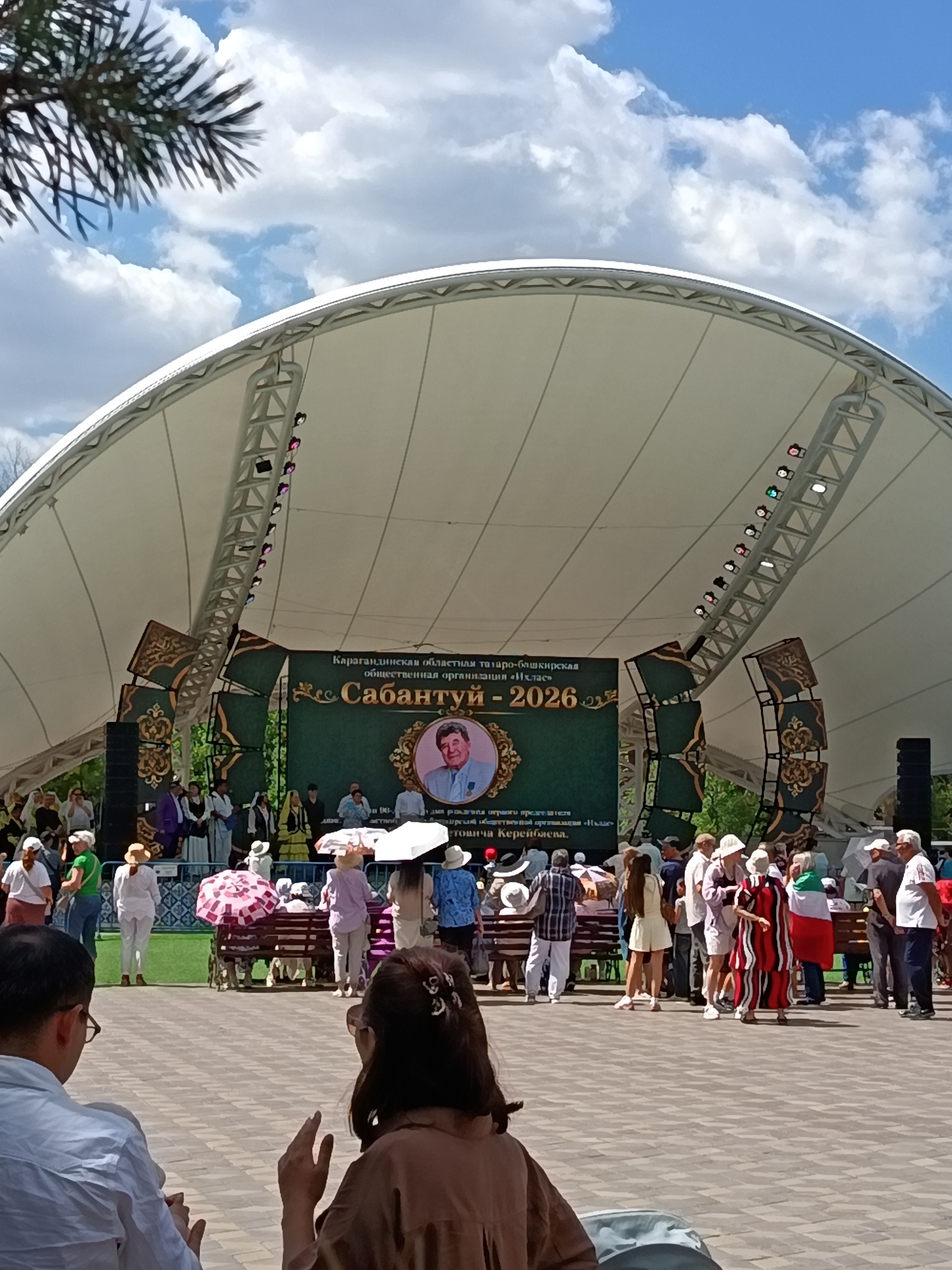 In the city of Qarağandı, Kazakhstan, an outdoor stage is set for an event under a large, white, arched canopy. The stage backdrop features a green banner with ornate golden detailing and text in Cyrillic script, along with a portrait of a man in a framed oval. A crowd of people, some dressed in traditional attire and others in casual wear, are gathered in front of and around the stage. Several individuals are holding umbrellas, suggesting sunny weather. Among the attendees, some are standing, while others are seated on benches or in the foreground. The ground is paved with patterned tiles. The overall scene depicts a community gathering or celebration.