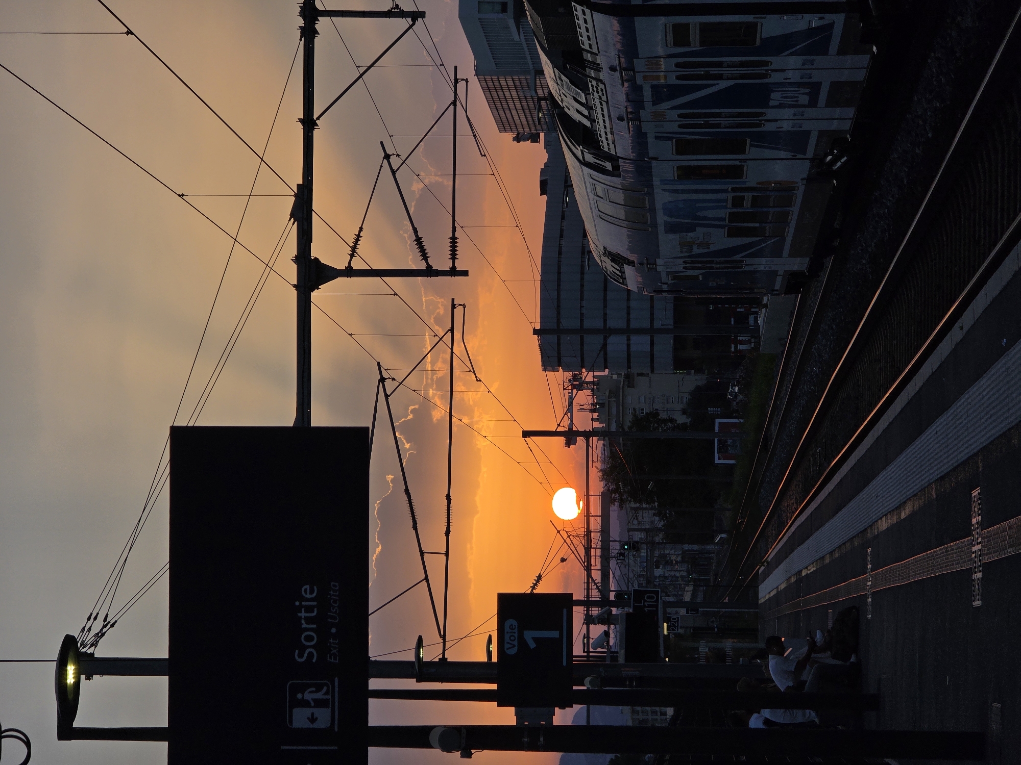 This image depicts a train station platform at sunset in Nice, France. The sky is a vibrant orange and yellow, with some clouds visible. A train is stopped at the platform, and several people are waiting. A large "Sortie" (Exit) sign is visible in the foreground, along with a sign indicating "Voie 1" (Track 1). The train is modern, with blue and white markings. Overhead, numerous electrical wires for the train are visible. The platform itself appears to be made of concrete with yellow safety markings. The sun is low on the horizon, casting long shadows and illuminating the scene with a warm glow. In the background, buildings of varying heights and architectural styles can be seen, typical of an urban environment. The overall impression is one of a bustling urban transit hub during a beautiful natural event.