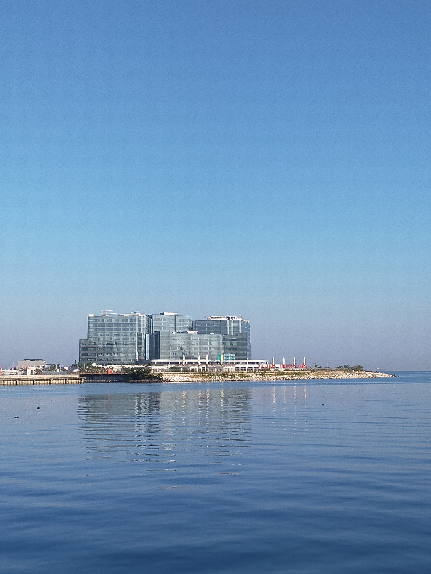 A bright, clear day in Makassar, Indonesia, showcases a modern waterfront development. The scene is dominated by a vast expanse of calm blue water reflecting the clear sky. In the distance, several large, multi-story glass buildings with sleek, contemporary architecture stand prominently on a strip of land bordering the water. The buildings appear to be commercial or office spaces, characterized by their reflective facades. A long, low-lying structure, possibly a promenade or a shopping area with white awnings, runs along the base of the buildings. The land is lined with a stone embankment and scattered with greenery. In the water, a few small, dark specks suggest birds or debris. The overall impression is one of urban development and leisure against a backdrop of serene natural beauty.