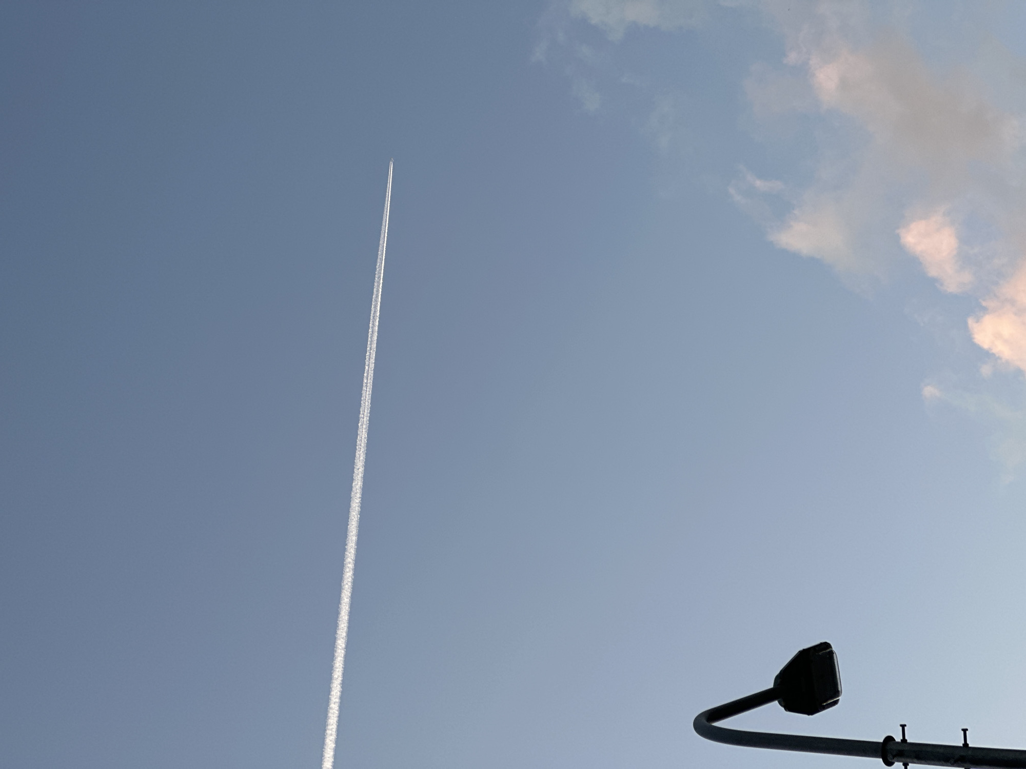 A silhouette of a streetlamp is visible in the lower left corner of the frame against a pale blue sky. A contrail from an airplane stretches horizontally across the upper portion of the sky. In the lower right quadrant, there are a few wispy clouds tinged with pink. The scene is likely a city in Kudrovo, Russia, given the context.