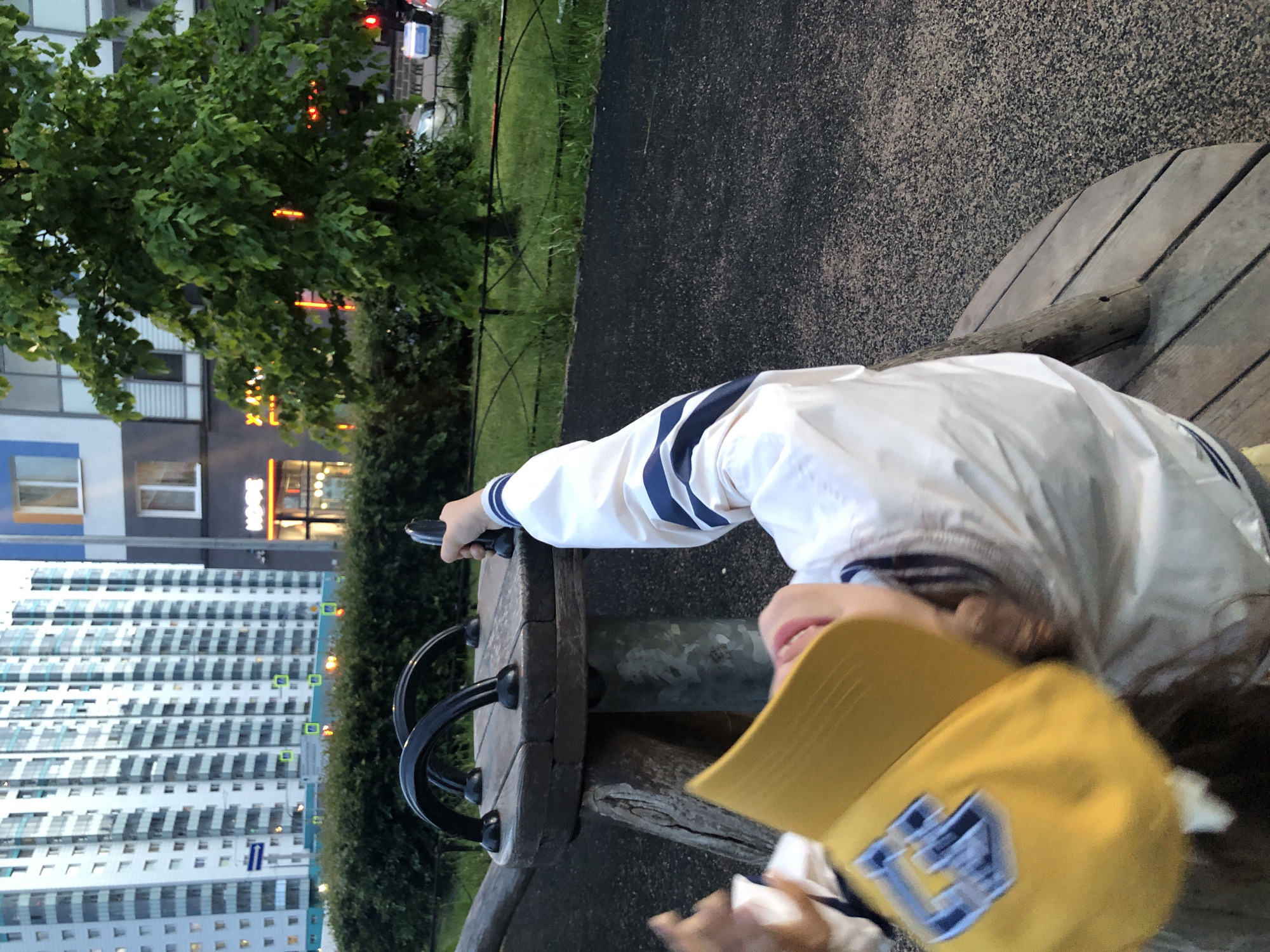 The image, taken from a slightly elevated perspective, captures a person in an urban park setting during dusk or early evening. The main subject, positioned in the lower right, wears a white jacket with navy blue stripes on the sleeves and a bright yellow baseball cap featuring a stylized blue letter, possibly "L," and other shapes. They are leaning on a rustic wooden structure, which appears to be part of a playground or outdoor seating, equipped with black metal handles. Their right arm is extended, and they seem to be pointing an object, possibly a smartphone, or simply their finger, towards the upper left of the frame. A slight smile is visible on their upturned face.
The immediate foreground includes a textured asphalt path and a strip of green grass, bordered by a black metal fence. In the background, a dense canopy of green trees occupies the left side. Beyond the trees, several multi-story buildings dominate the horizon, characteristic of modern residential developments in a city. One building with a dark facade prominently displays a brightly lit red sign, which appears to spell "HOCK" or "HOSE." Further in the distance, a tall, light-colored high-rise apartment building with numerous balconies stands under a dimming sky. The artificial lights from the buildings indicate the transition from day to night.