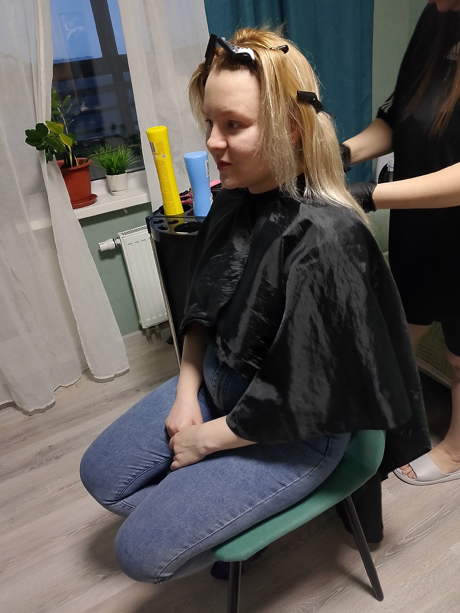 A woman is receiving a hair treatment in what appears to be an indoor setting. She is seated on a green stool, wearing blue jeans and a black cape to protect her clothing. Her blonde hair is partially clipped up with black hair clips. A person, presumably a hairstylist, stands behind her, wearing black gloves and a black shirt, tending to her hair. In the background, there is a window with sheer white curtains, a radiator, and a windowsill with potted plants. There is also a black stand with yellow and blue bottles. The floor is a light-colored wood laminate. The scene takes place in a city with a population under 700,000 people.