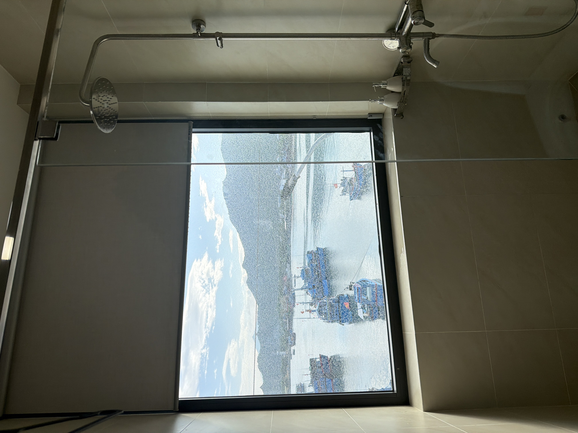The image captures a modern shower area featuring a large window that offers a picturesque outdoor view. The interior consists of light, neutral-toned tiles on the walls and ceiling. A sleek chrome shower system is mounted overhead and across the ceiling, including a large rain showerhead on the left and various controls, possibly a handheld shower, and wall-mounted dispensers on the right. A glass panel or screen, reflecting the interior, separates the shower space.
The focal point is the dark-framed window, which is noticeably wet, suggesting recent or ongoing showering activity. Through the water-splashed glass, a vibrant daytime landscape is visible. A calm body of water, likely a bay or a large river, stretches into the distance, dotted with several distinctive blue boats, which appear to be fishing or aquaculture vessels. In the background, majestic green mountains or hills rise under a bright blue sky with scattered white clouds. A grey roller blind is partially lowered on the left side of the window.
The scene depicts a sunny day, with ample natural light illuminating the outdoor environment. The wet window provides a unique visual filter to the stunning vista. No people or visible text are present. The combination of modern bathroom amenities with a striking natural backdrop of mountains, water, and boats is consistent with coastal regions, such as Ninh Chu Ward, Vietnam.
