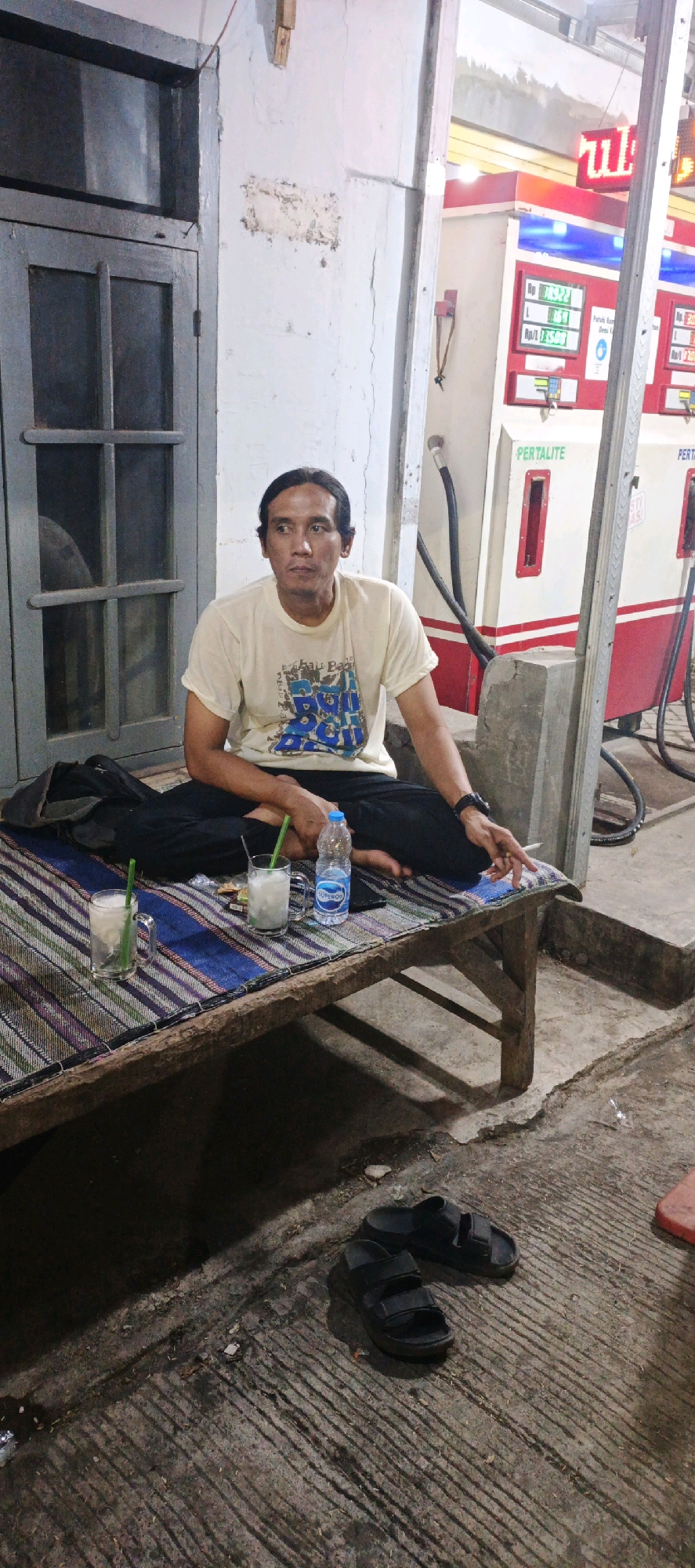 A man is sitting cross-legged on a woven mat placed on a wooden platform in front of a white building with gray trim. He is wearing a light-colored t-shirt with a graphic print and dark pants. In front of him on the mat are two glasses of a white beverage with green straws, a bottle of water, and a mobile phone. He is holding a cigarette in his right hand. Behind him, a gray door with glass panes is visible. To the right of the man are two gas pumps with red and white detailing, with signage that reads "PERTALITE." The ground in front of the platform is concrete with visible grooves, and a pair of black sandals are lying on it. The scene appears to be outdoors, possibly at a gas station in Kasiyan, Indonesia.