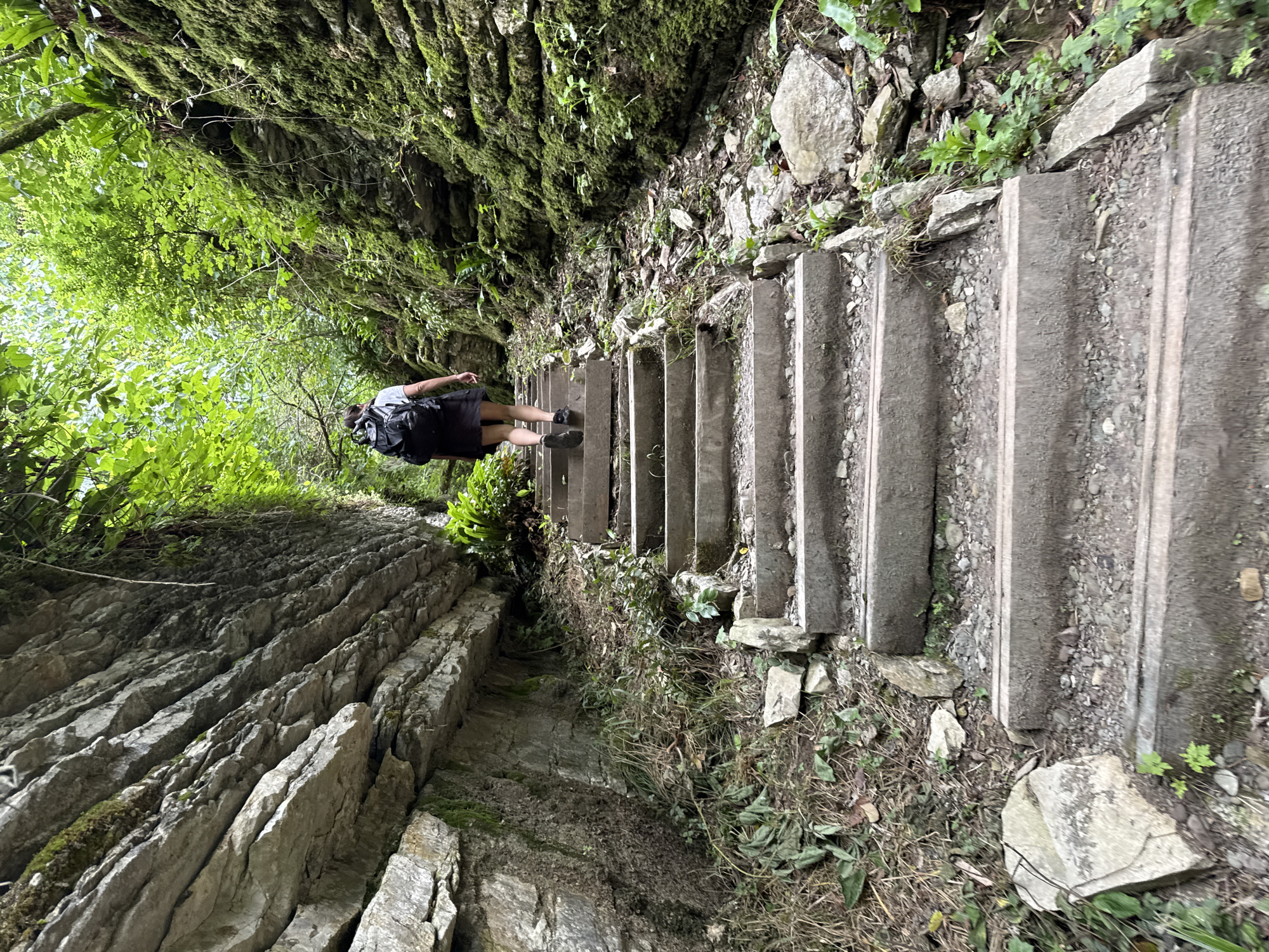 A person with a backpack walks up a stone staircase in a narrow, forested gorge. The staircase is made of concrete slabs and is flanked by moss-covered rock walls on either side. Lush green vegetation grows on the walls and along the sides of the path. The lighting suggests it is daytime, possibly overcast. This scene appears to be a natural attraction or a hiking trail, potentially within the city of Khleborob, Russia, given the prompt's context.