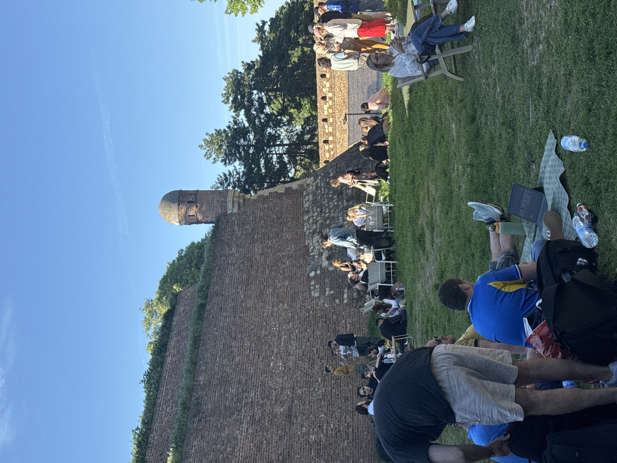 The image depicts a scene at the Belgrade Fortress in Belgrade, Serbia. A large brick wall runs vertically on the left side of the image, with a tower structure at the top of the wall. On the right side of the image is a grassy area where numerous people are sitting, some on chairs and others on the grass. Some individuals are talking to each other. A few items are placed on the grass including water bottles, a laptop, and a blanket.